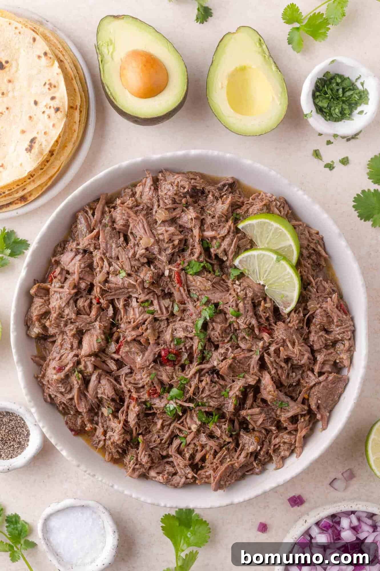 Effortless Shredded Barbacoa 2 A large bowl of shredded slow cooker barbacoa beef garnished with fresh lime slices, surrounded by tortillas, avocados, and bowls of toppings.