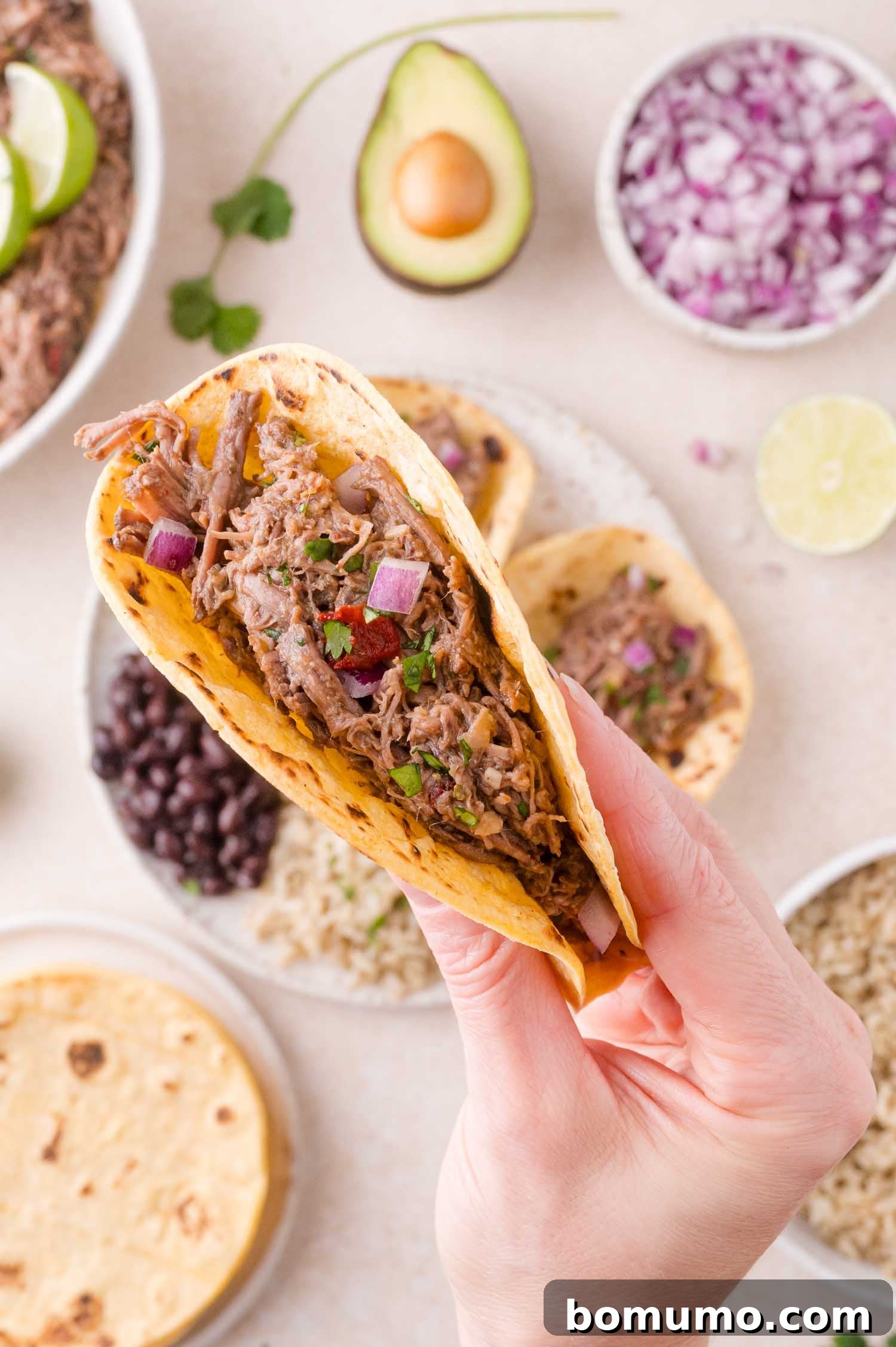 Effortless Shredded Barbacoa 14 A hand holding a barbacoa taco filled with shredded beef, onions, and cilantro over a table with various toppings.