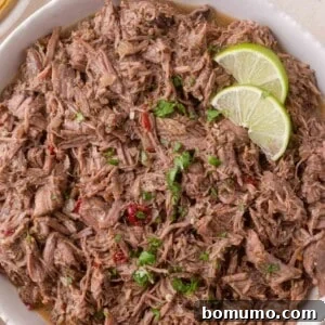 Effortless Shredded Barbacoa 15 A square image of shredded beef barbacoa in a white bowl, garnished with cilantro and lime.