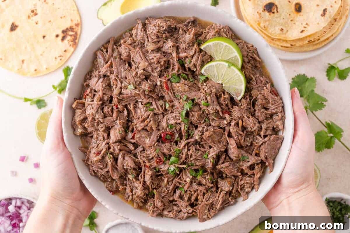 Effortless Shredded Barbacoa 3 A person holding a bowl of freshly made slow cooker barbacoa, with tortillas and other fresh ingredients blurred in the background.