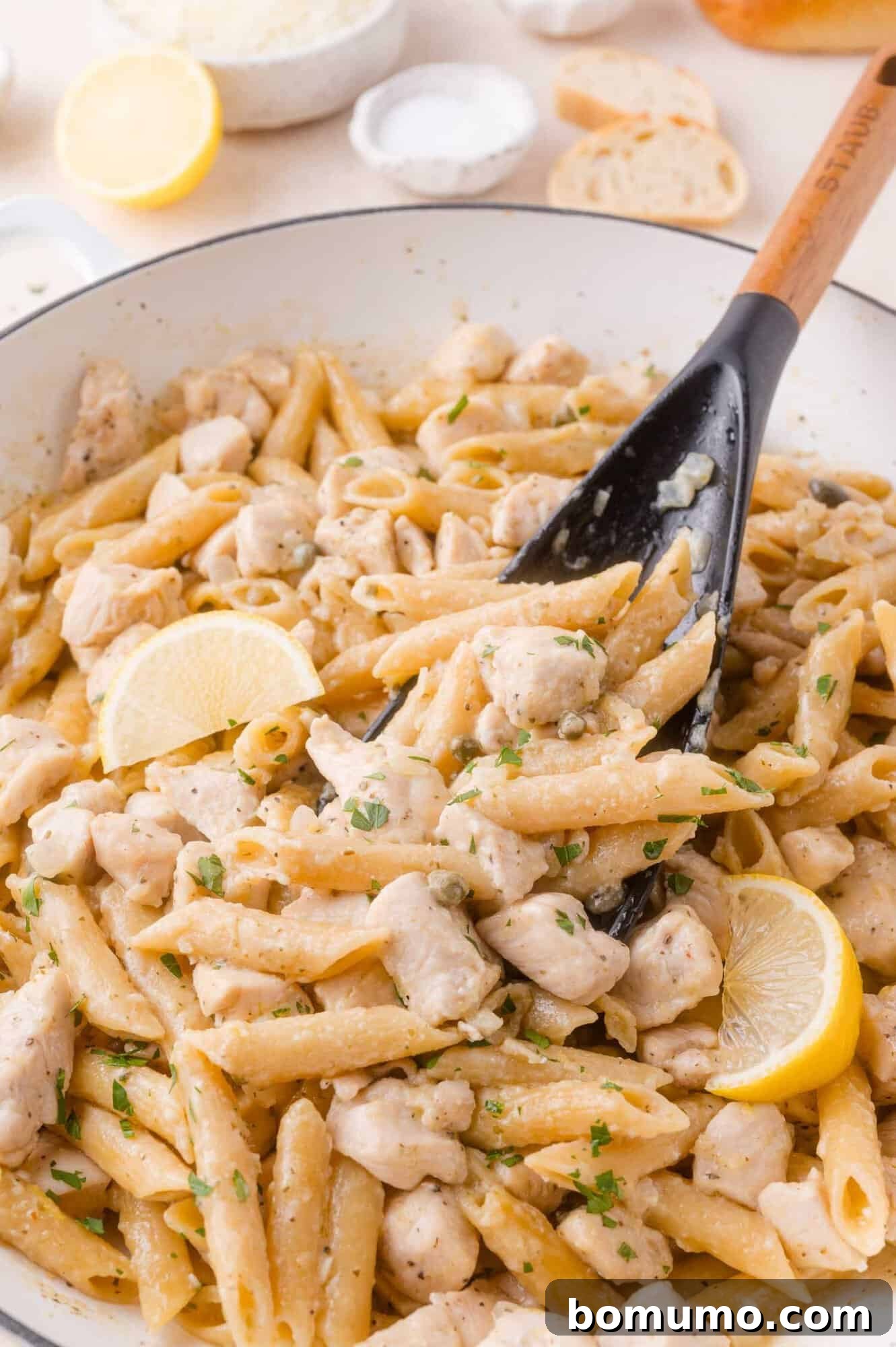 A pan of creamy chicken piccata pasta with chunks of chicken, garnished with lemon slices and parsley, being stirred with a black spoon.