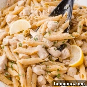 Penne chicken piccata pasta with diced chicken, lemon wedges, and herbs, mixed in a creamy sauce, served in a white dish with a black serving spoon.