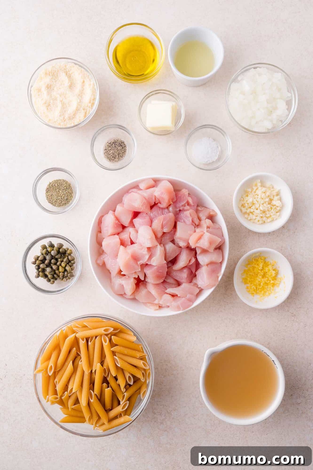 Overhead view of ingredients in small bowls on a countertop, including diced chicken, uncooked penne pasta, chopped onion, minced garlic, capers, broth, butter, oil, seasonings, and cheese.