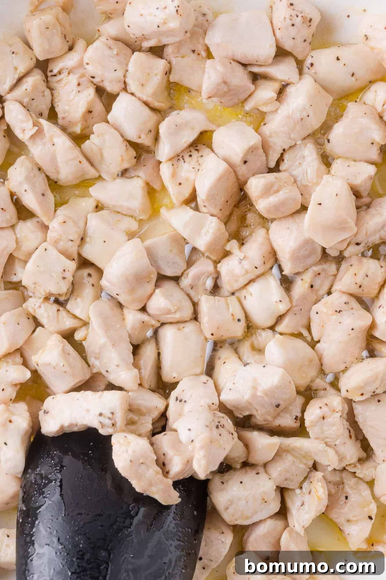 Cubed pieces of chicken breast are being cooked in a pan, with a black spatula stirring the lightly browned and seasoned meat.