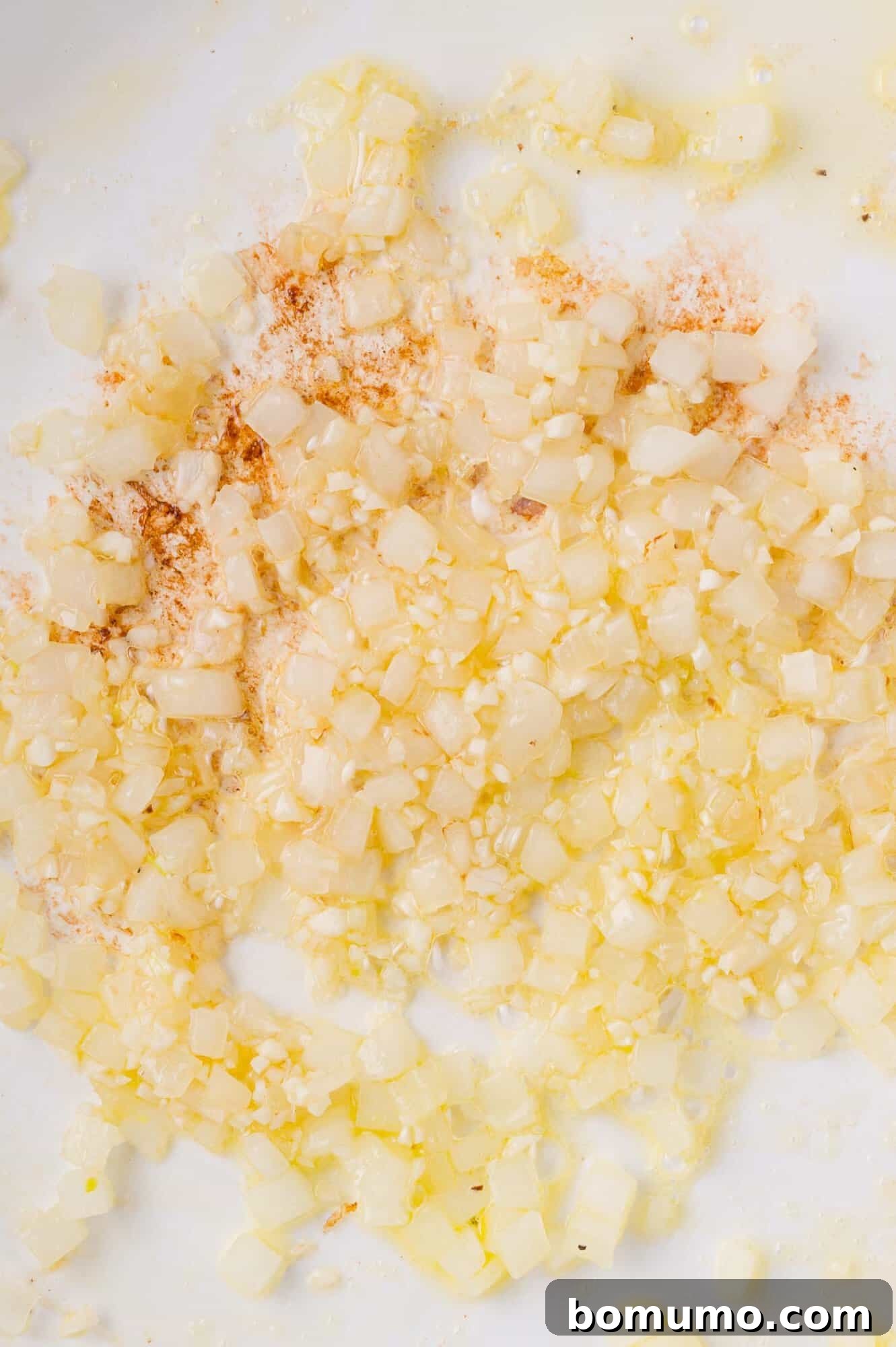 Chopped onions and minced garlic sautéing in oil with a sprinkle of spices on a white surface.