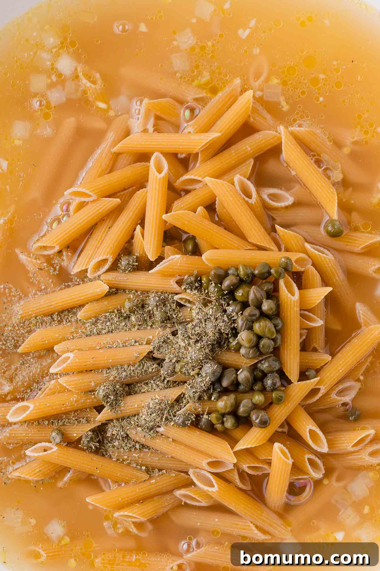 Uncooked penne pasta, capers, herbs, and spices in a broth or liquid, prepared for cooking.
