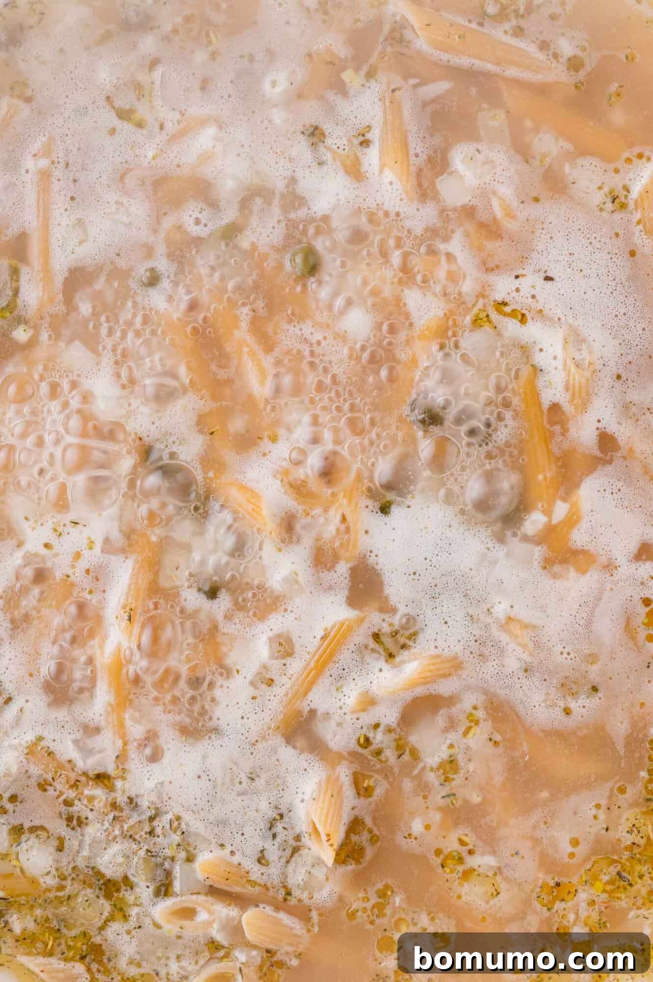 Close-up of pasta cooking in boiling water with bubbles, floating foam, and visible herbs and pasta pieces.