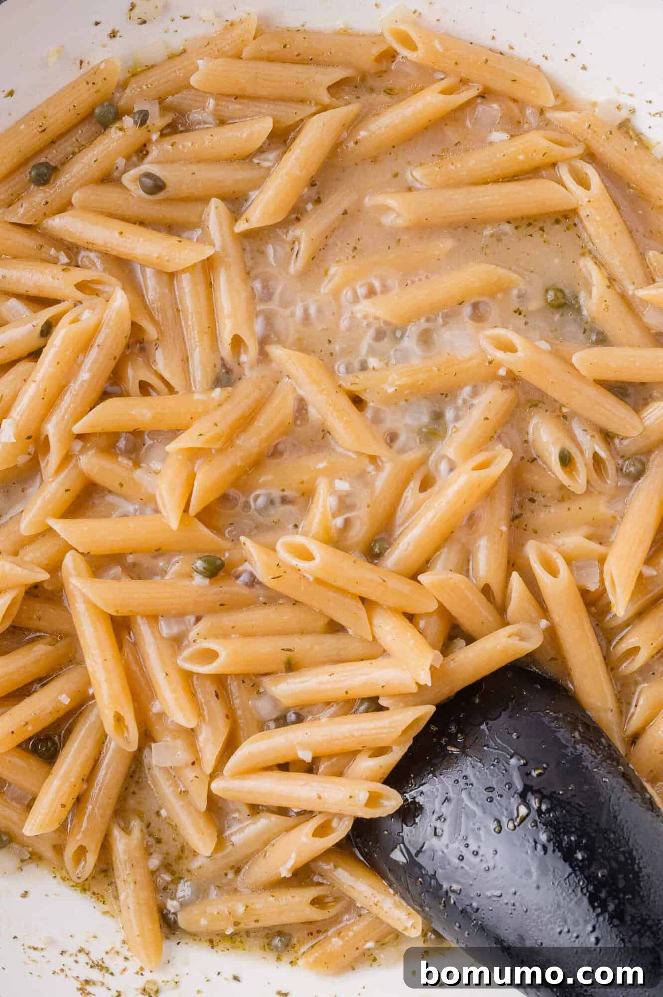 Cooked penne pasta in a creamy sauce with visible herbs and capers, stirred with a black serving spoon in a white pan.