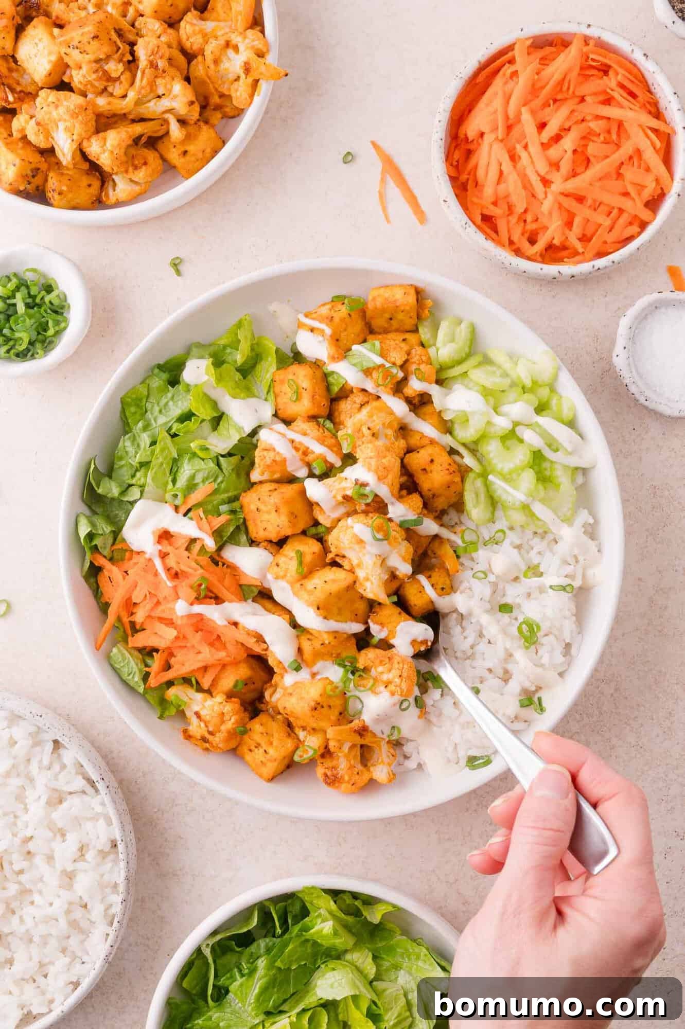 A bowl of rice, lettuce, shredded carrots, seasoned tofu pieces, and ranch dressing, with a hand holding a fork. Surrounding bowls contain tofu, carrots, rice, and lettuce.