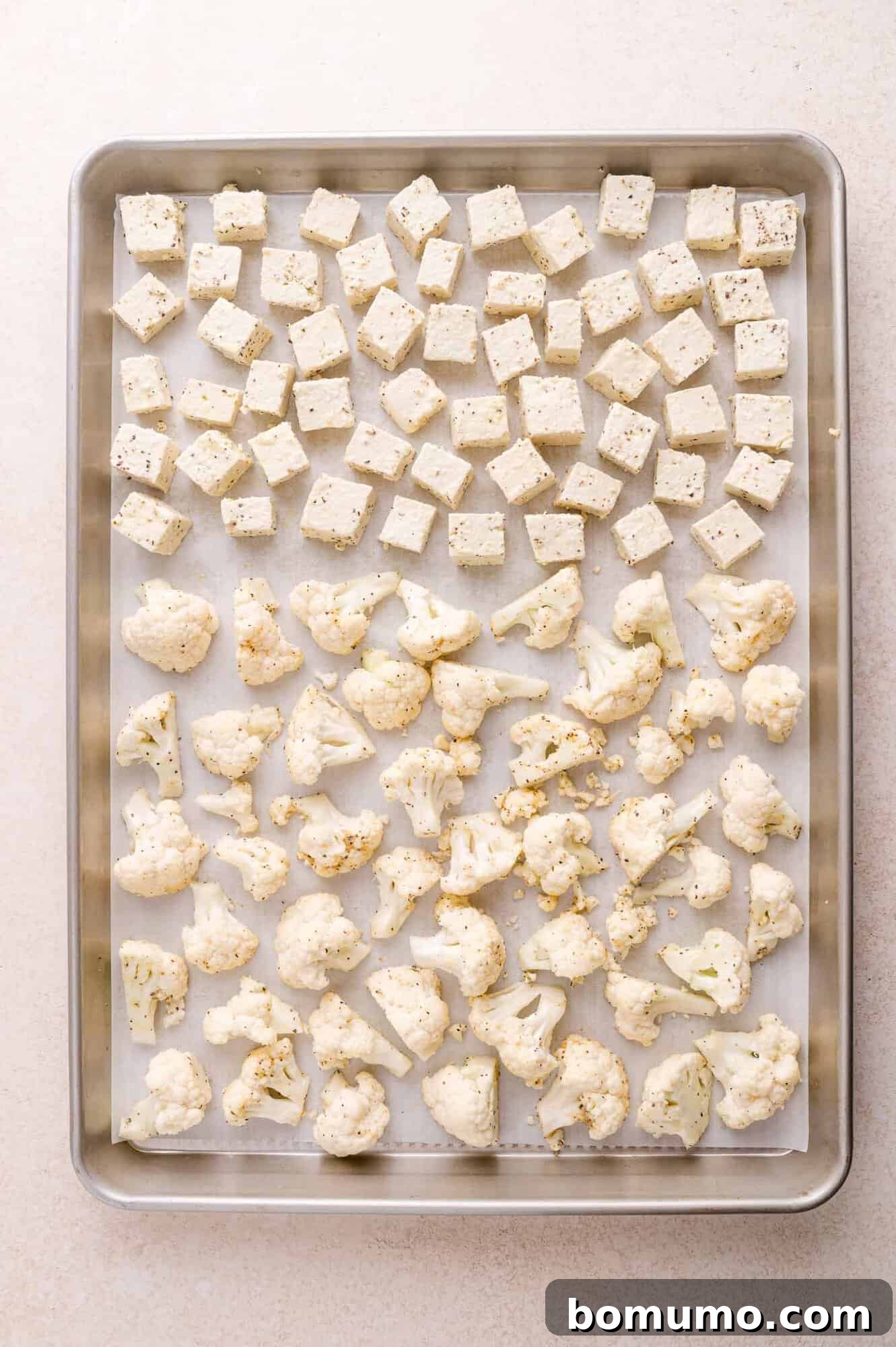 A baking sheet with evenly spaced cubes of tofu and cauliflower florets, both seasoned, on parchment paper.