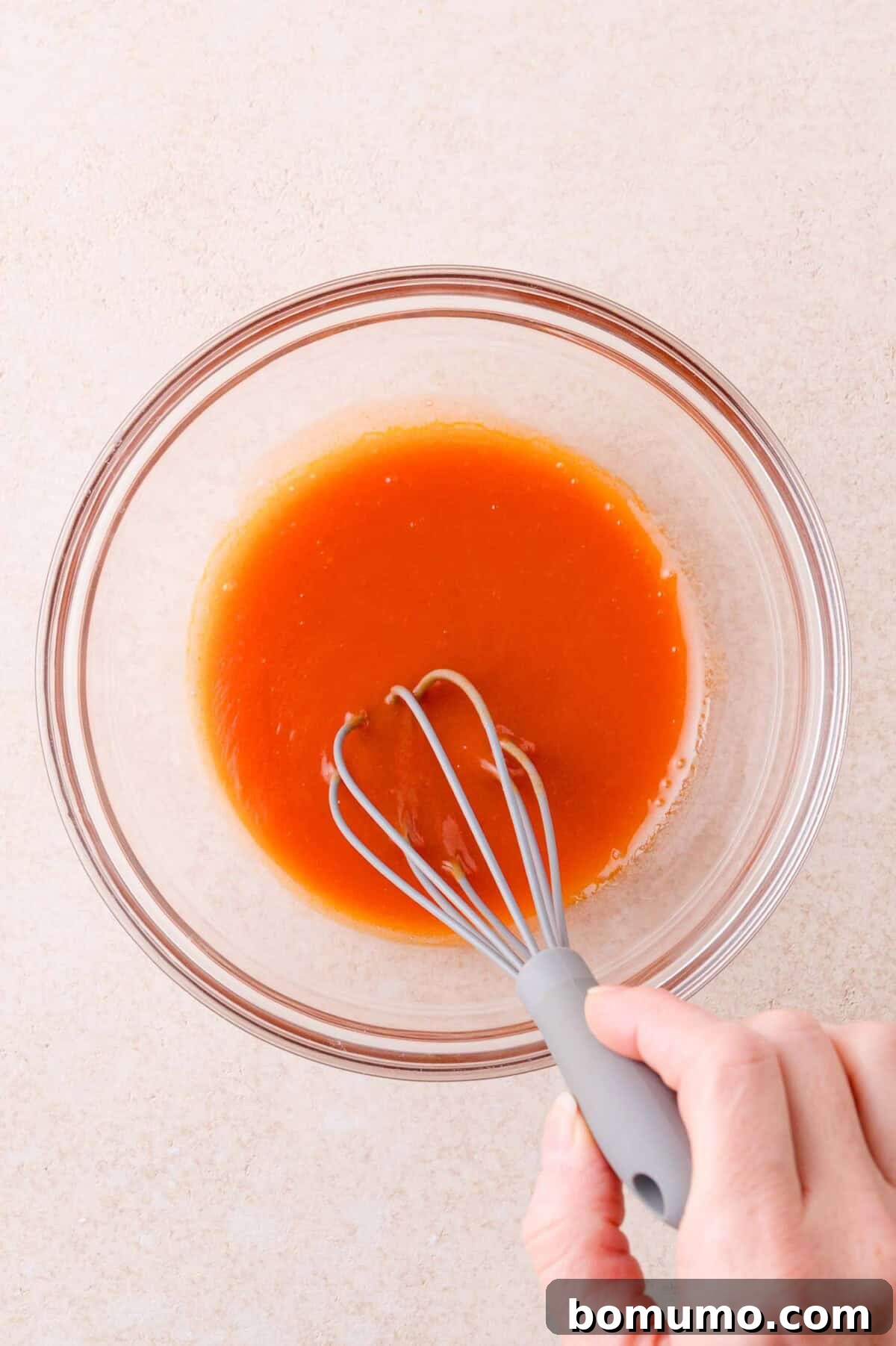 A hand holding a whisk mixes an orange liquid in a clear glass bowl on a light-colored surface.