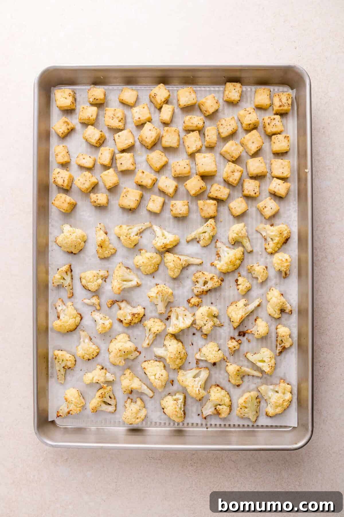 Baking sheet lined with parchment paper holding evenly spaced roasted cauliflower florets on the bottom half and roasted tofu cubes on the top half.