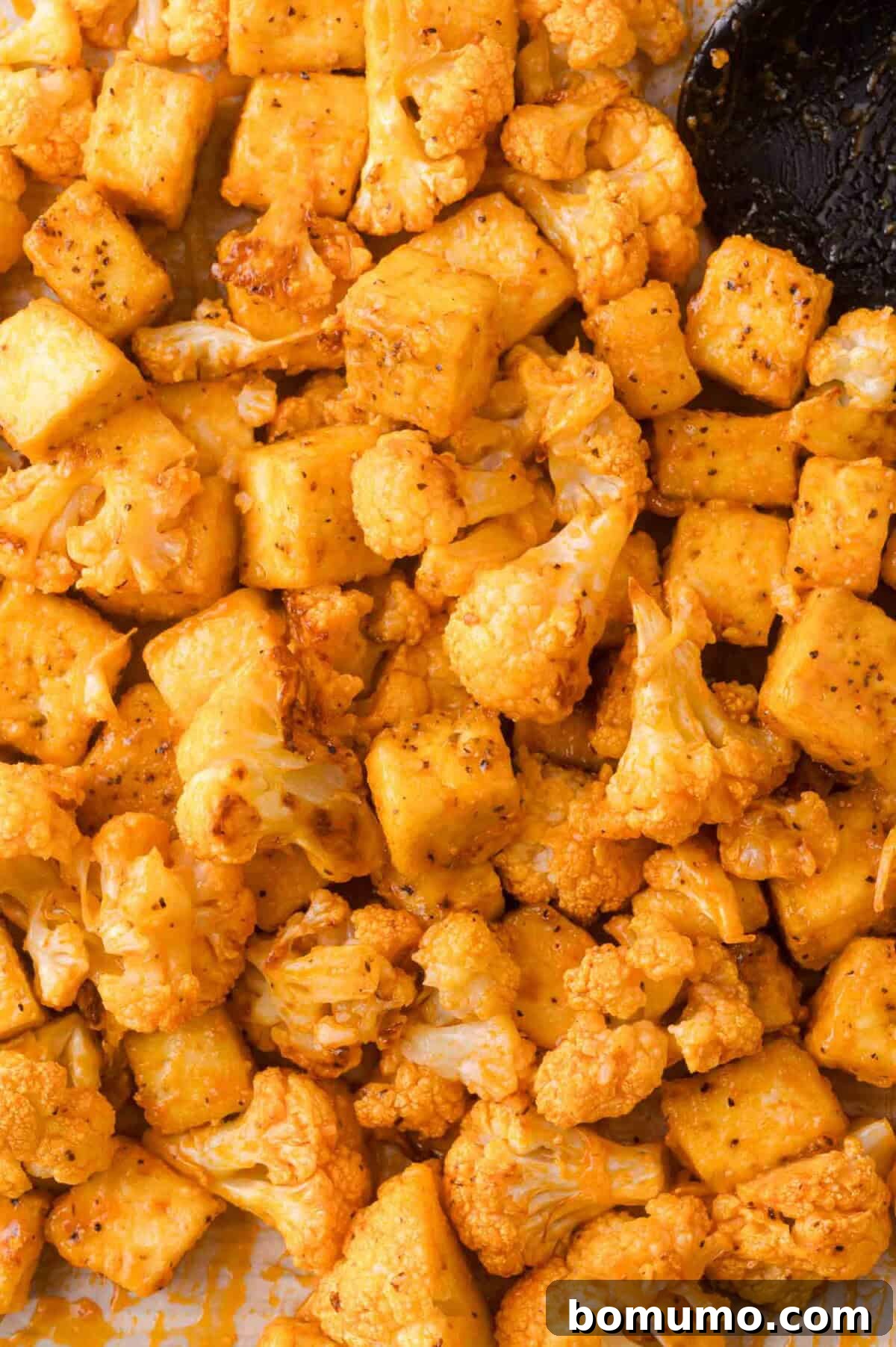 Roasted cauliflower florets and tofu cubes coated in spices, arranged on a baking sheet with a black spatula partially visible.