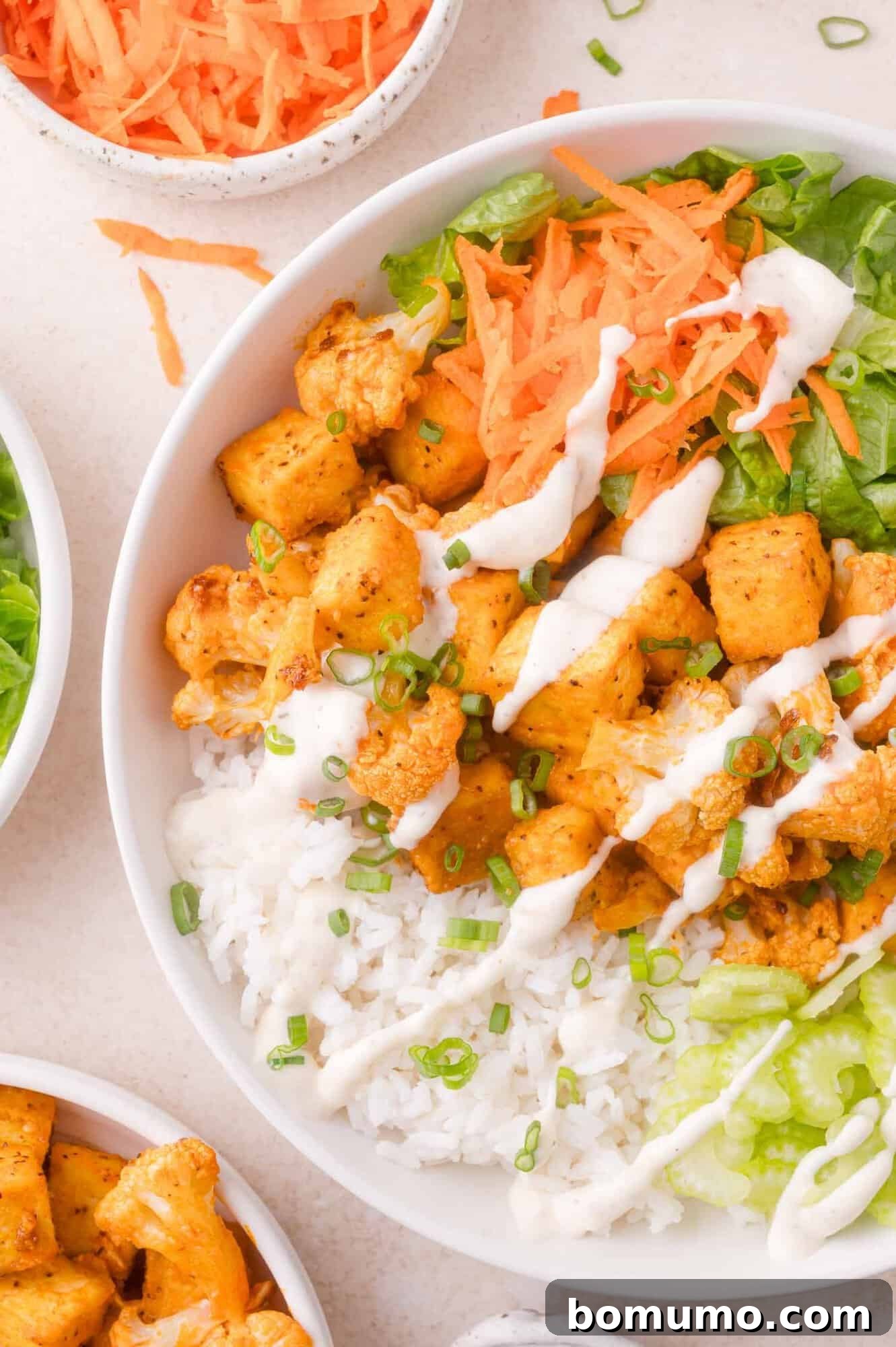 A bowl filled with white rice, lettuce, shredded carrots, seasoned tofu and cauliflower, and drizzled with a ranch dressing, garnished with chopped green onions.
