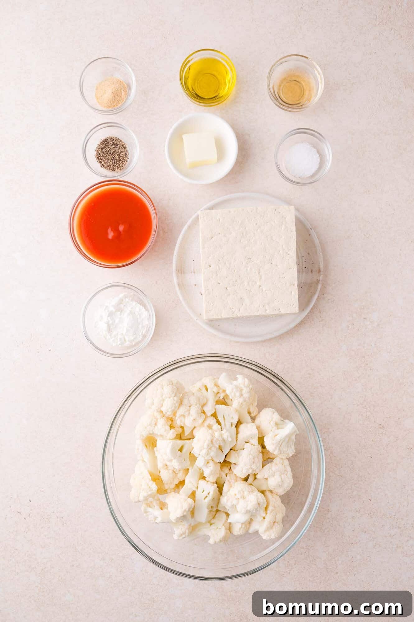 A glass bowl of cauliflower florets, a block of tofu, and small bowls containing hot sauce, oil, butter, vinegar, cornstarch, garlic powder, pepper, and salt arranged on a light surface.
