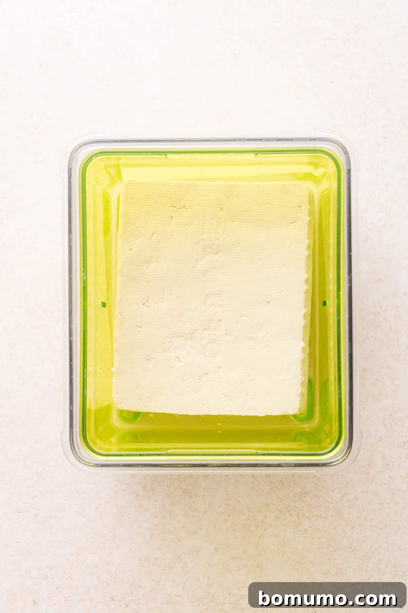 A block of tofu sits in a transparent container with a green base, filled with liquid, viewed from above on a light surface.