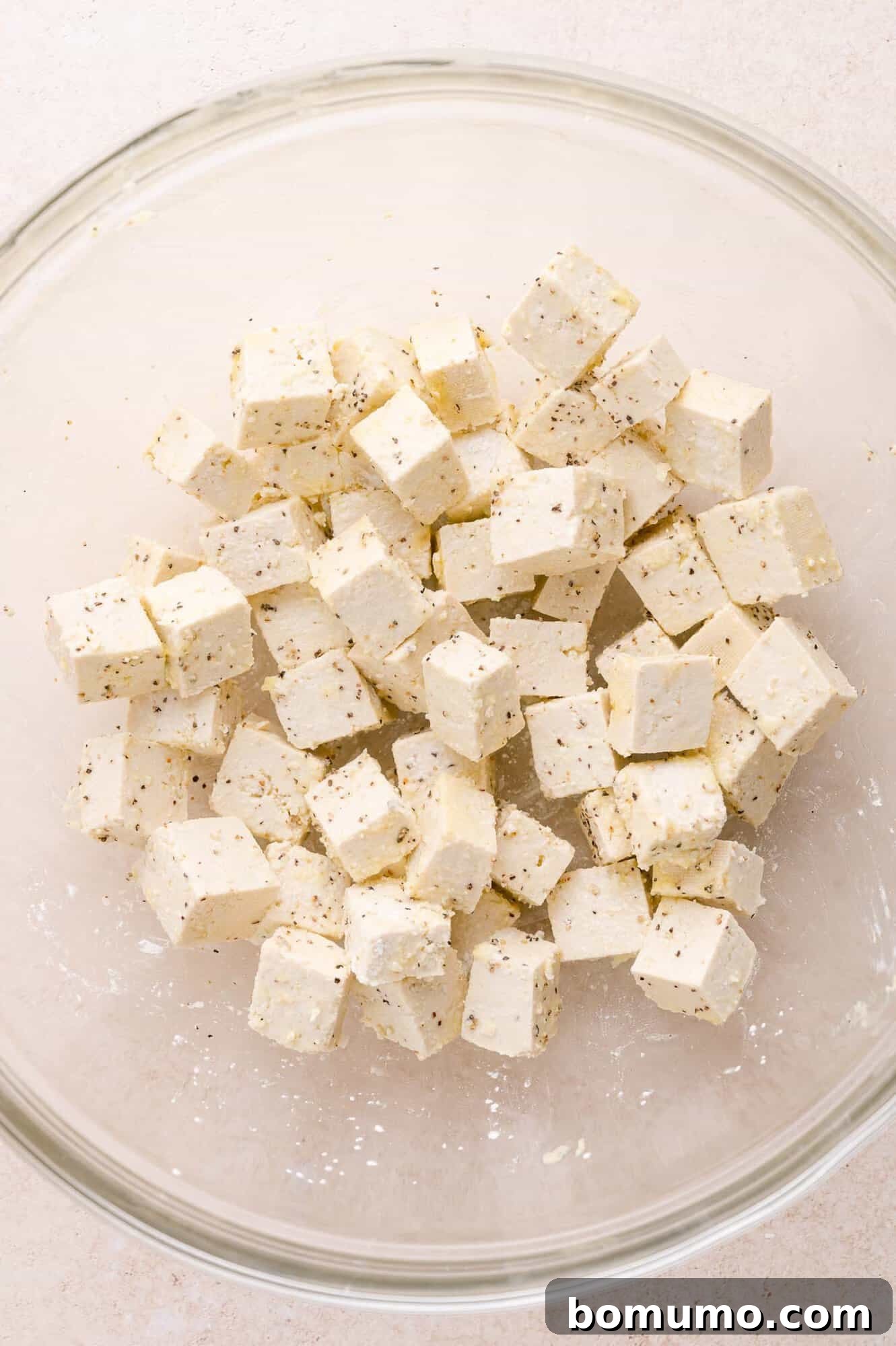 Cubed tofu seasoned with black pepper and spices in a clear glass bowl on a light-colored surface.