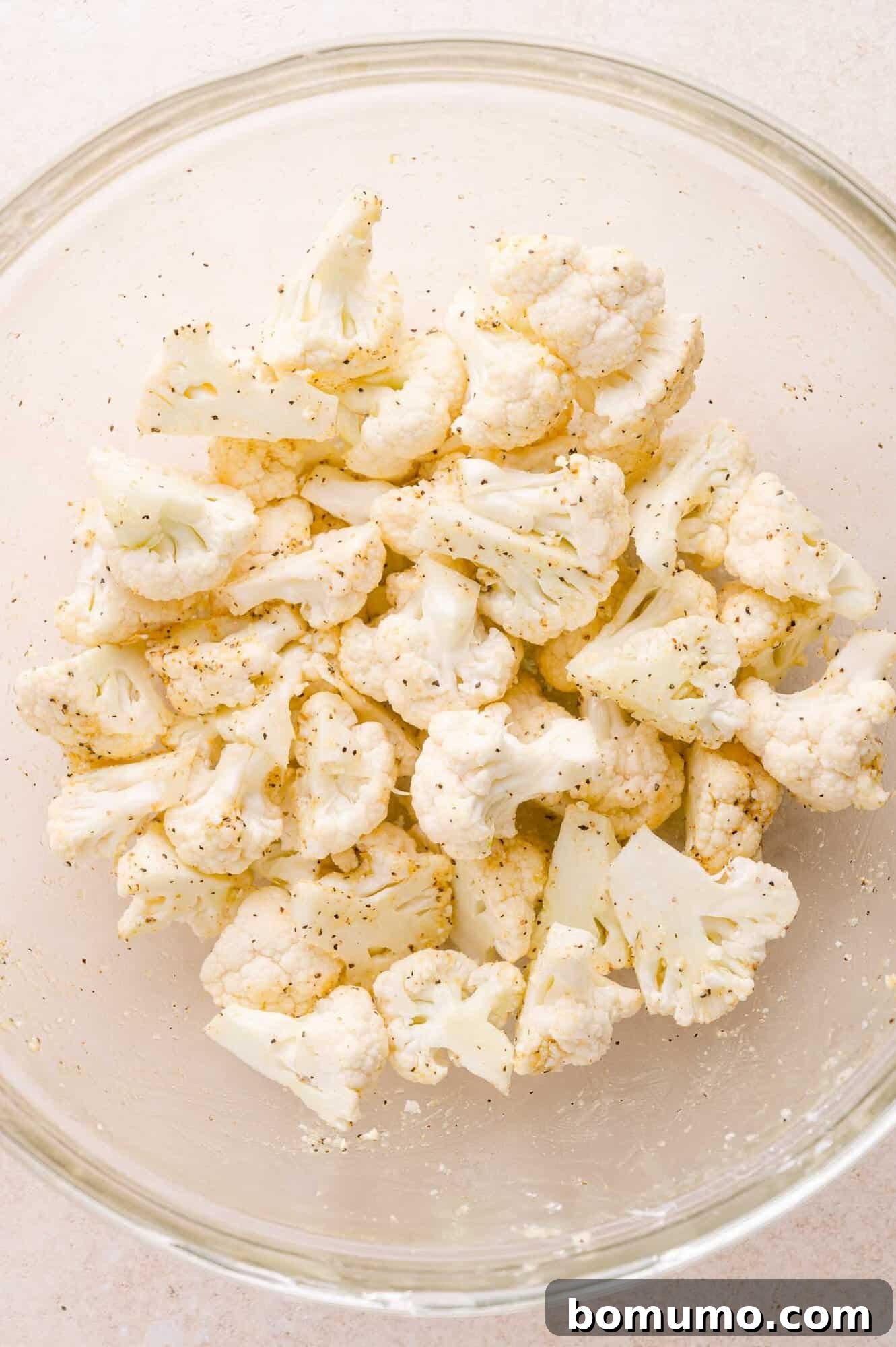 Raw cauliflower florets in a glass bowl, seasoned with salt, pepper, and other spices.
