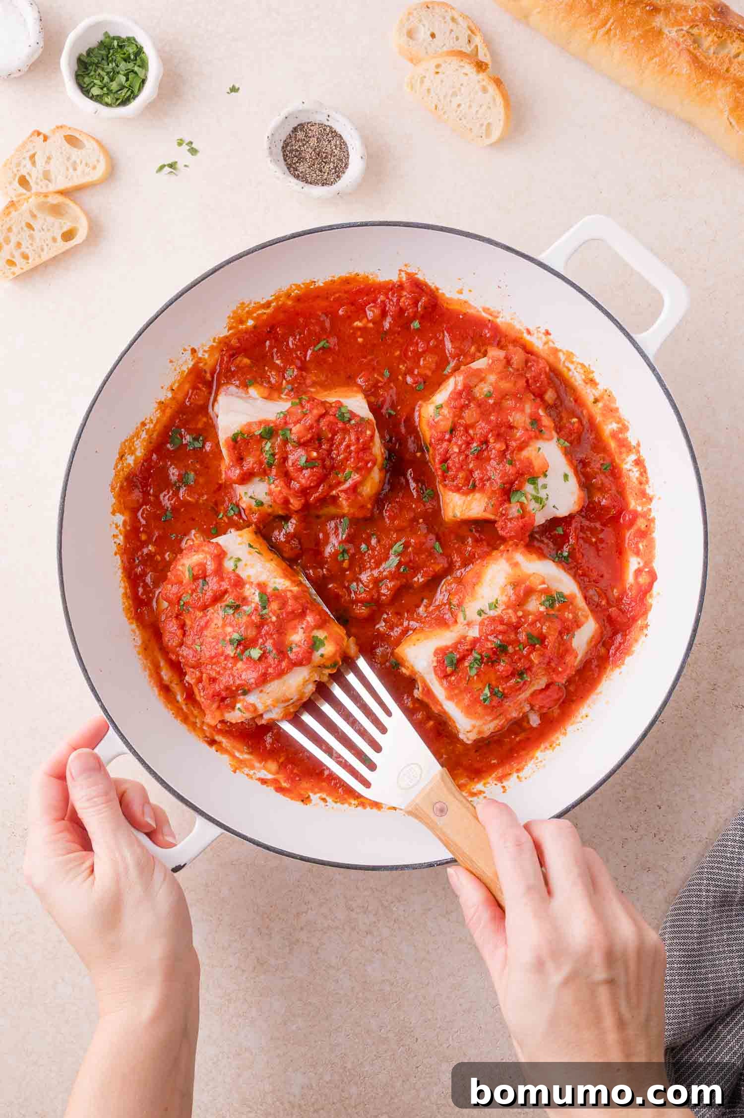A person serves white cod fillets in tomato sauce from a white pan, surrounded by small bowls of herbs, pepper, and slices of baguette.