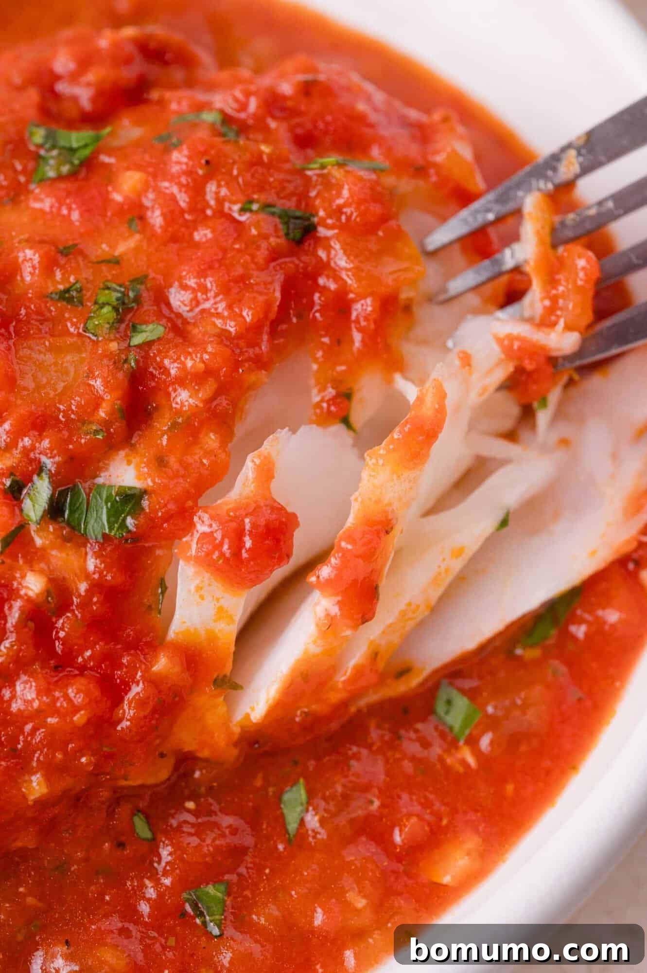 A close-up of a flaky cod fillet topped with chunky tomato sauce and garnished with chopped herbs, being pulled apart with a fork.