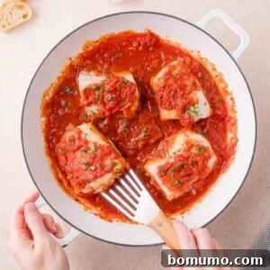 Four pieces of cod are cooked in a skillet with chunky tomato sauce and garnished with herbs; a spatula is lifting one piece.