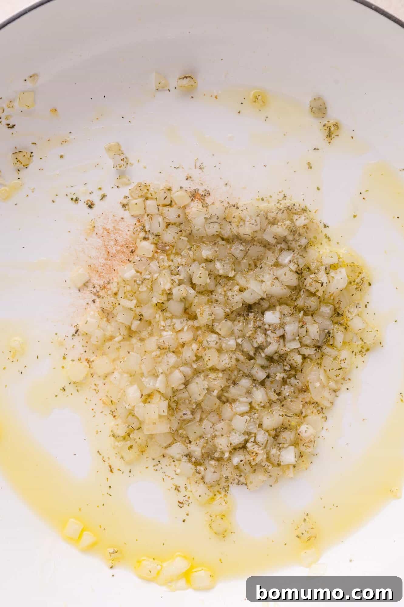 Diced onions sautéing in oil with herbs and spices in a white pan.