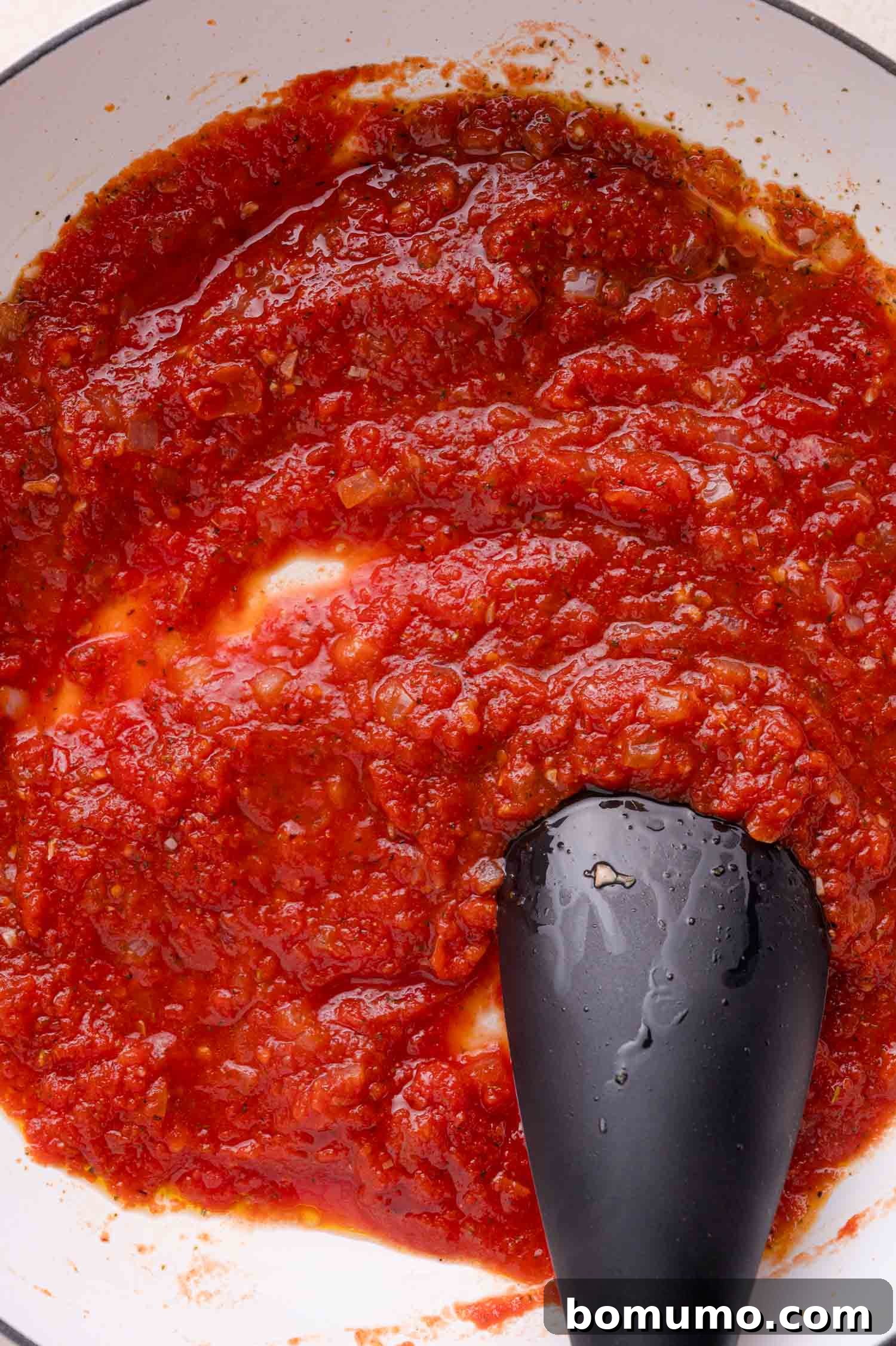 Tomato sauce being stirred in a white pan with a black silicone spatula.
