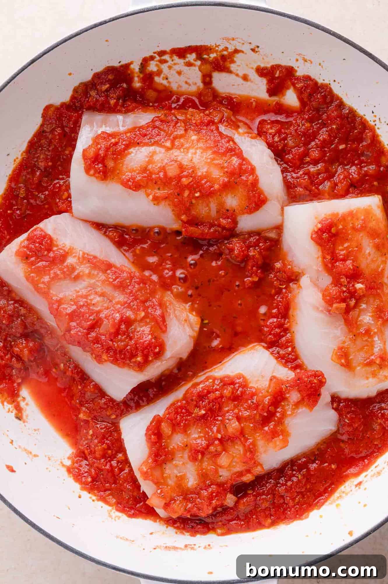 Four pieces of raw white cod in a pan, topped with chunky tomato sauce, ready to be cooked.
