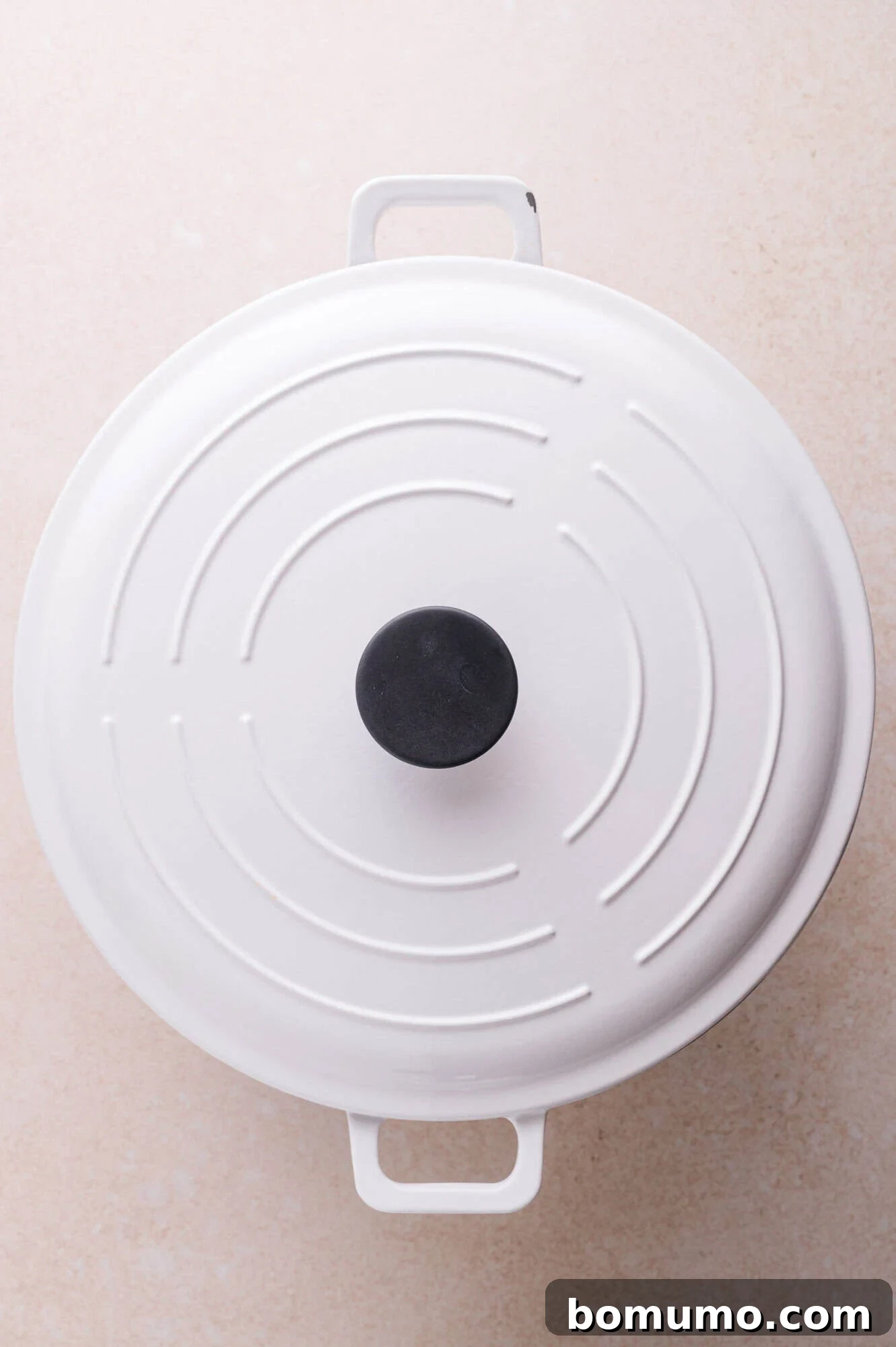 A white enameled cast iron Dutch oven with a black knob sits on a light-colored surface, viewed from above.