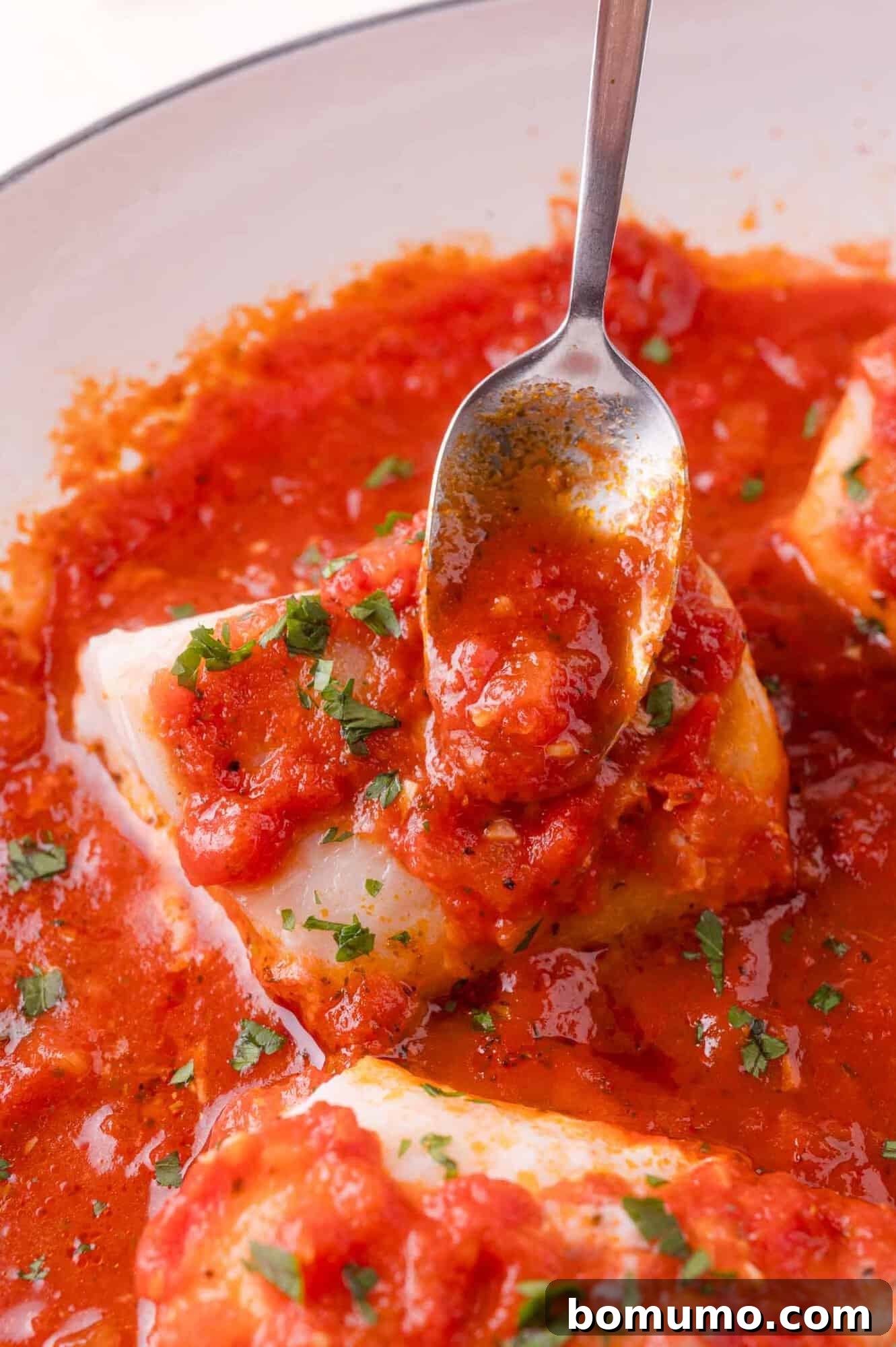 A spoon pours tomato sauce over a piece of white fish, garnished with chopped herbs, in a white dish.