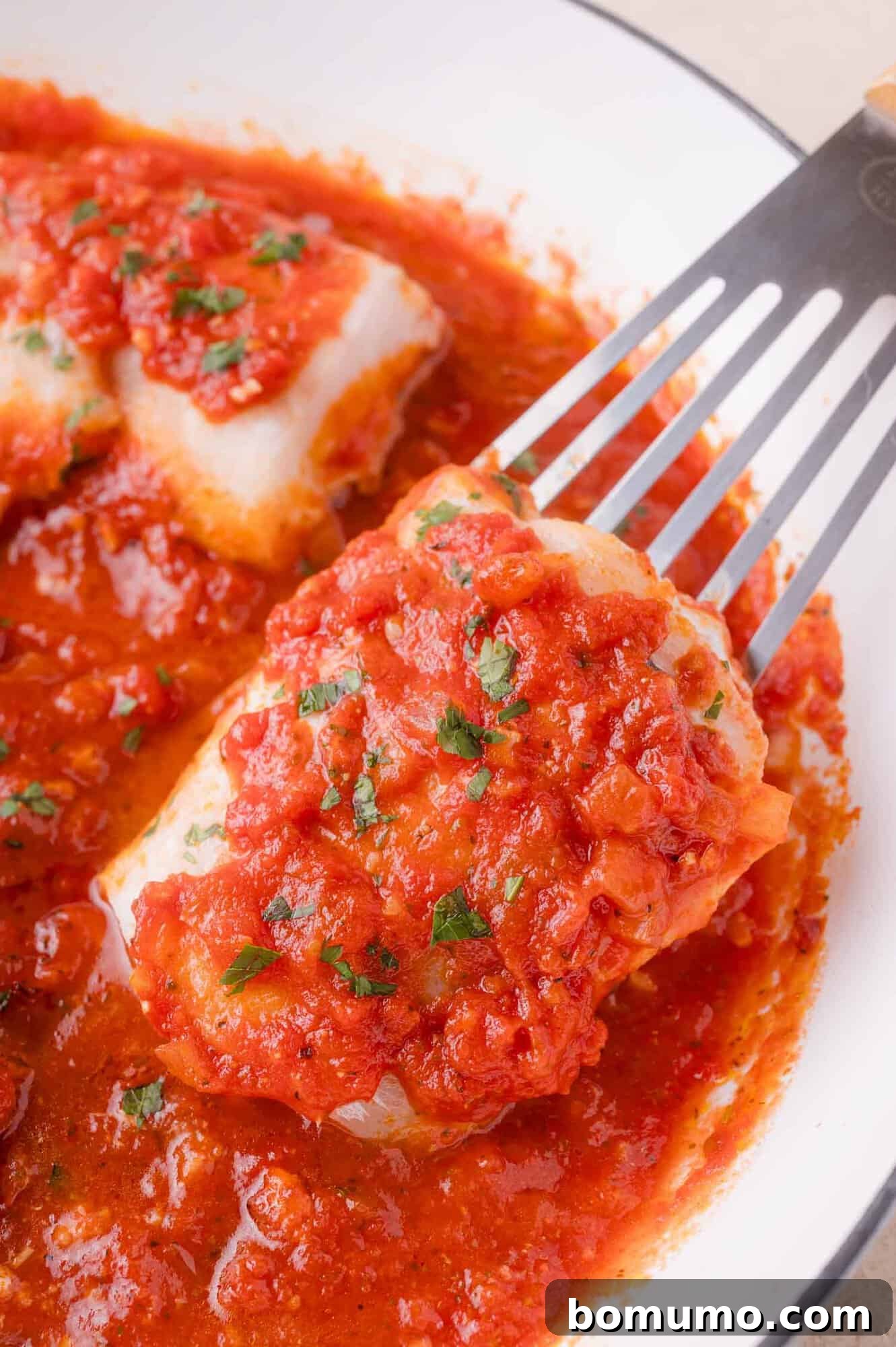 A piece of white fish topped with tomato sauce and herbs is lifted from a pan filled with tomato sauce using a metal spatula.