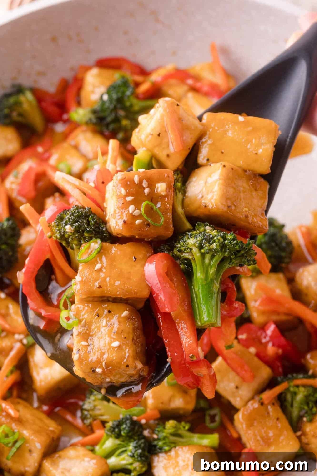 A close-up of a serving spoon holding tofu cubes, broccoli, red bell pepper, and carrot strips in a savory sauce.