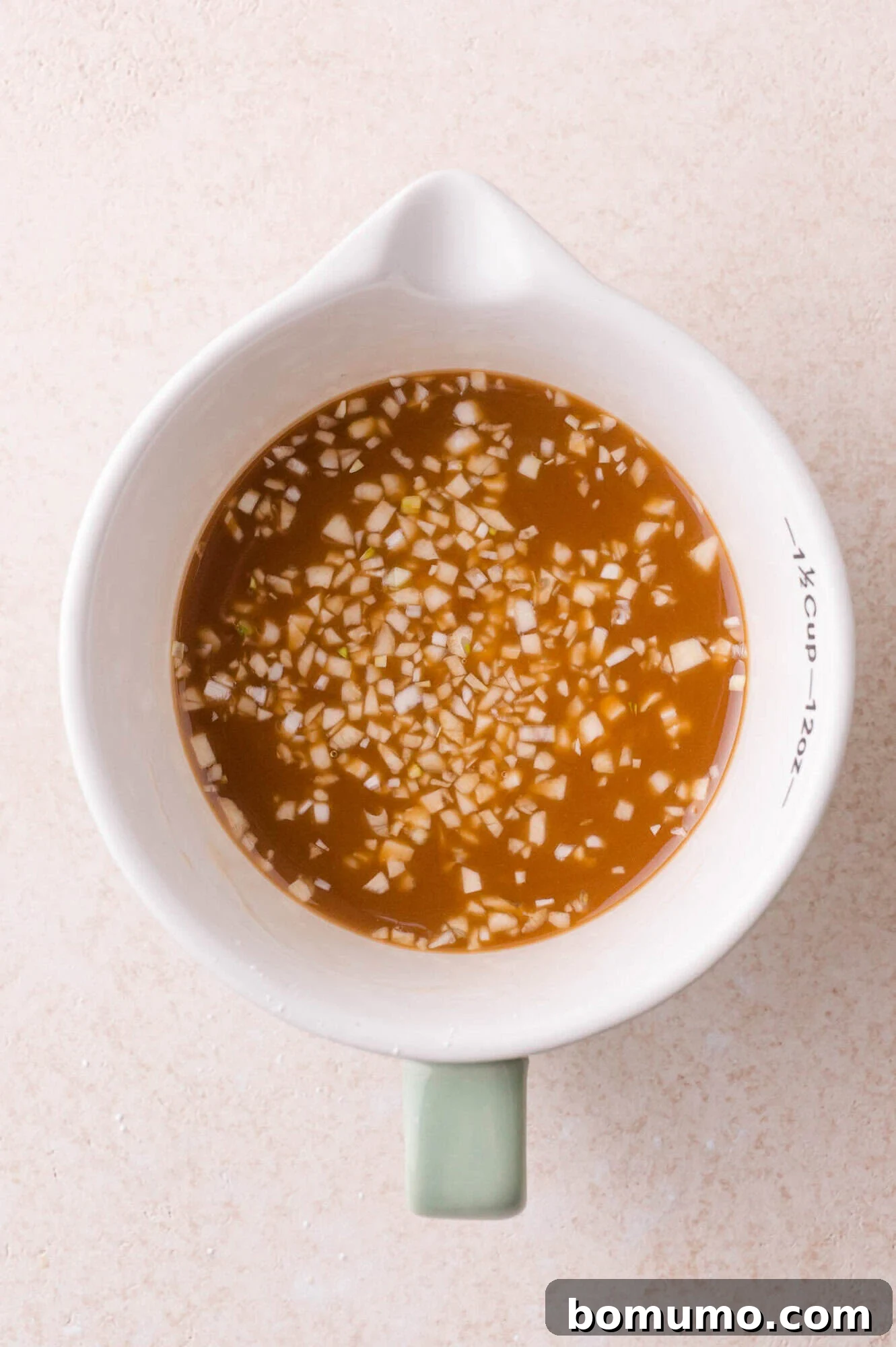 A white measuring cup filled with a brown liquid containing finely chopped garlic, placed on a light beige surface.