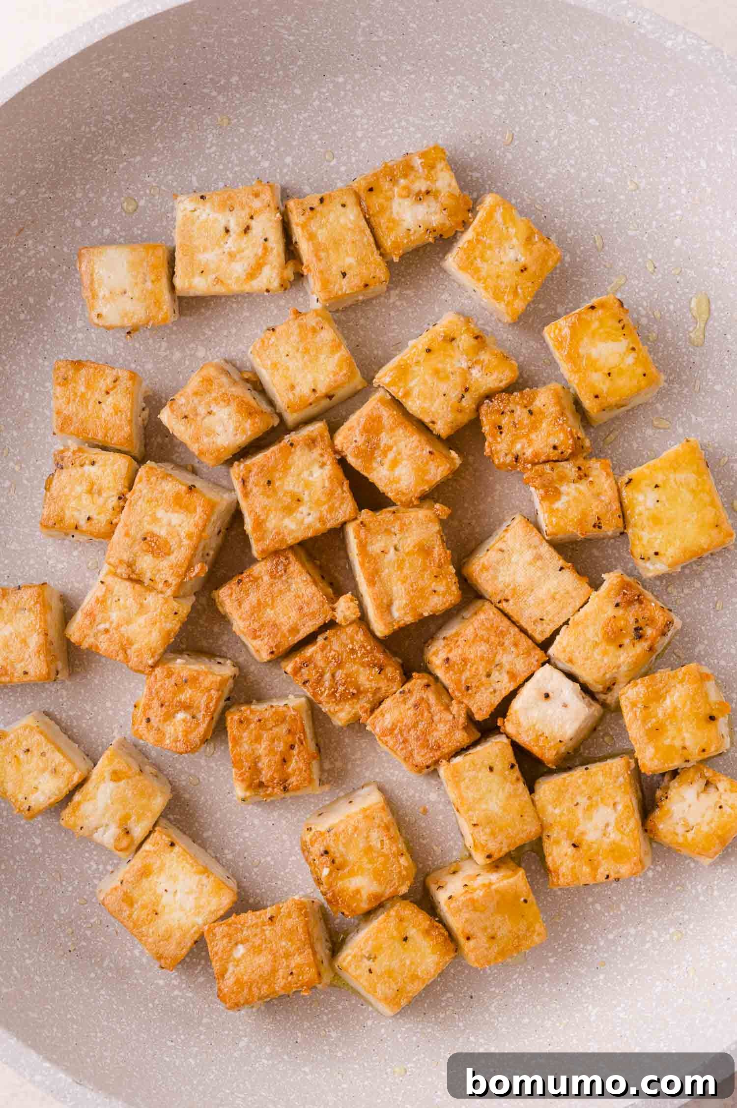 Cubed tofu is pan-fried to a golden brown in a light-colored skillet.