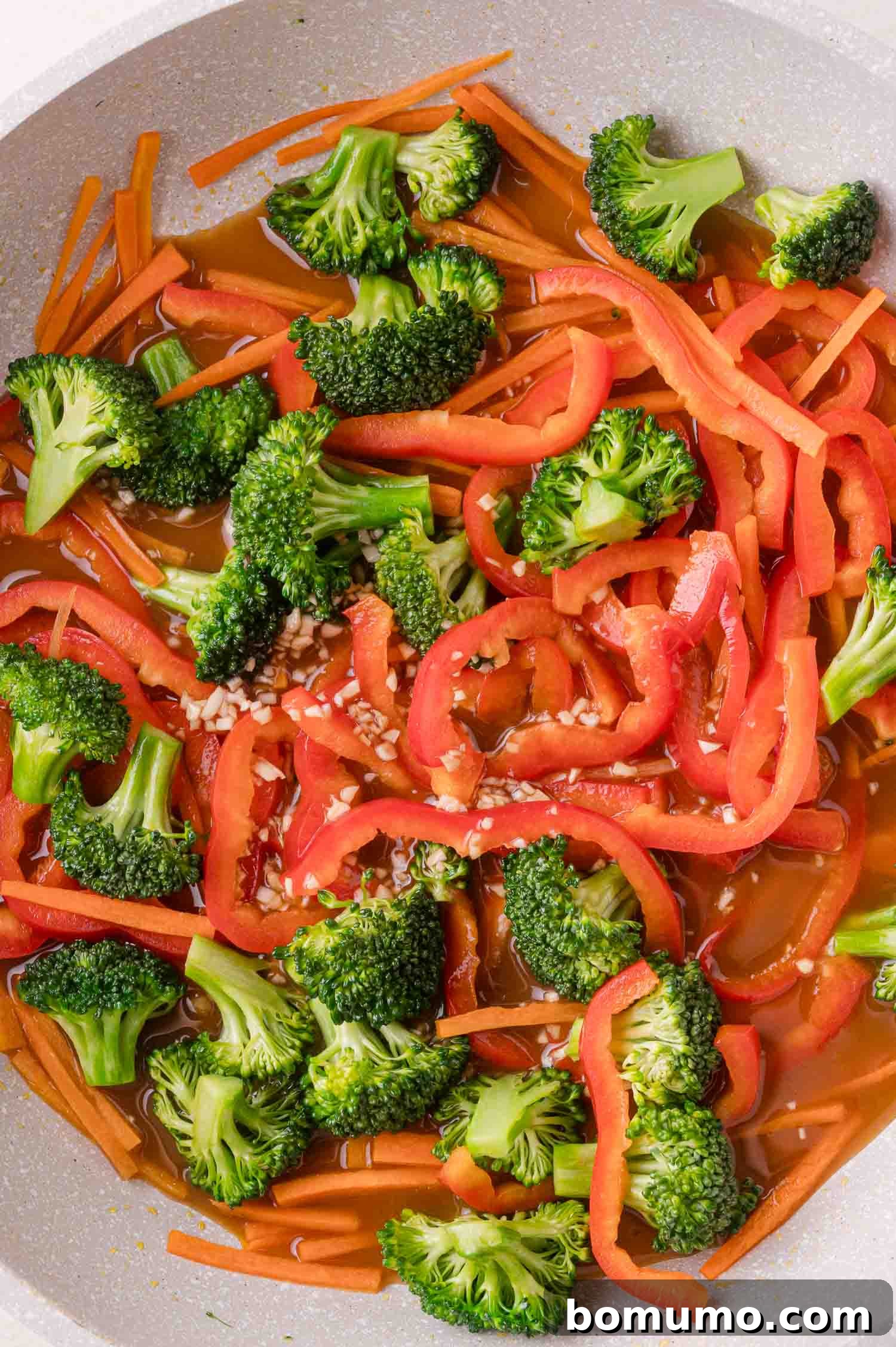 Broccoli florets, sliced red bell peppers, and carrots cooking in a pan with a brown sauce and minced garlic.