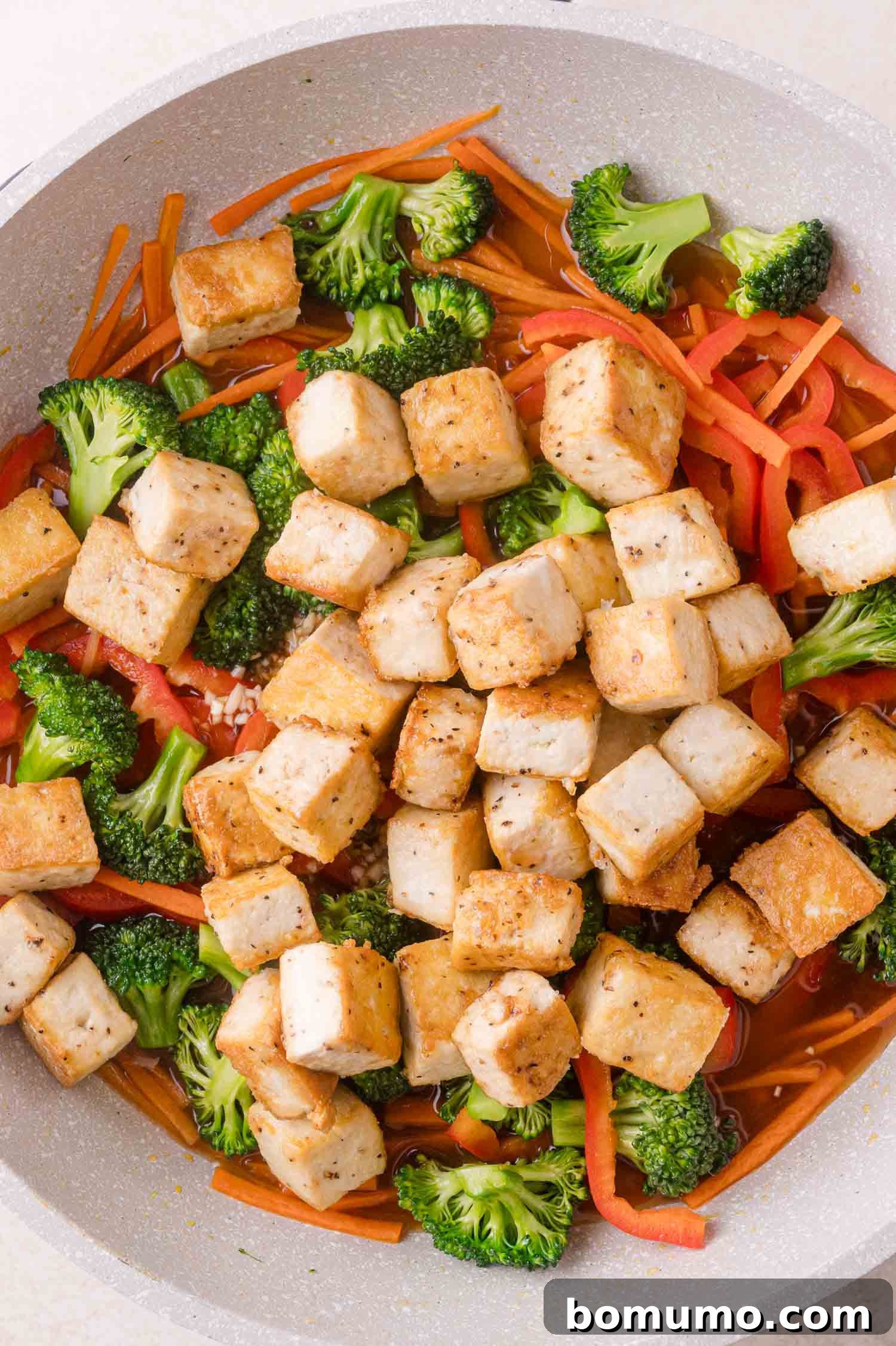 A pan filled with cooked tofu cubes, broccoli florets, sliced red bell pepper, and julienned carrots.