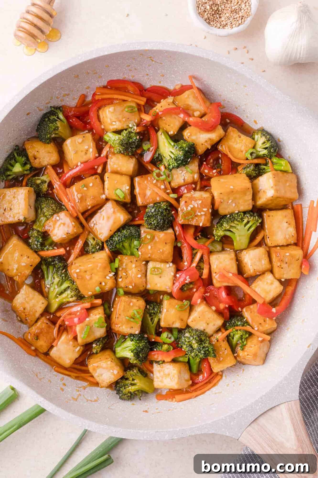 A stir-fry dish with cubed tofu, broccoli, red bell peppers, and carrots in a light sauce, topped with sesame seeds, served in a pan.