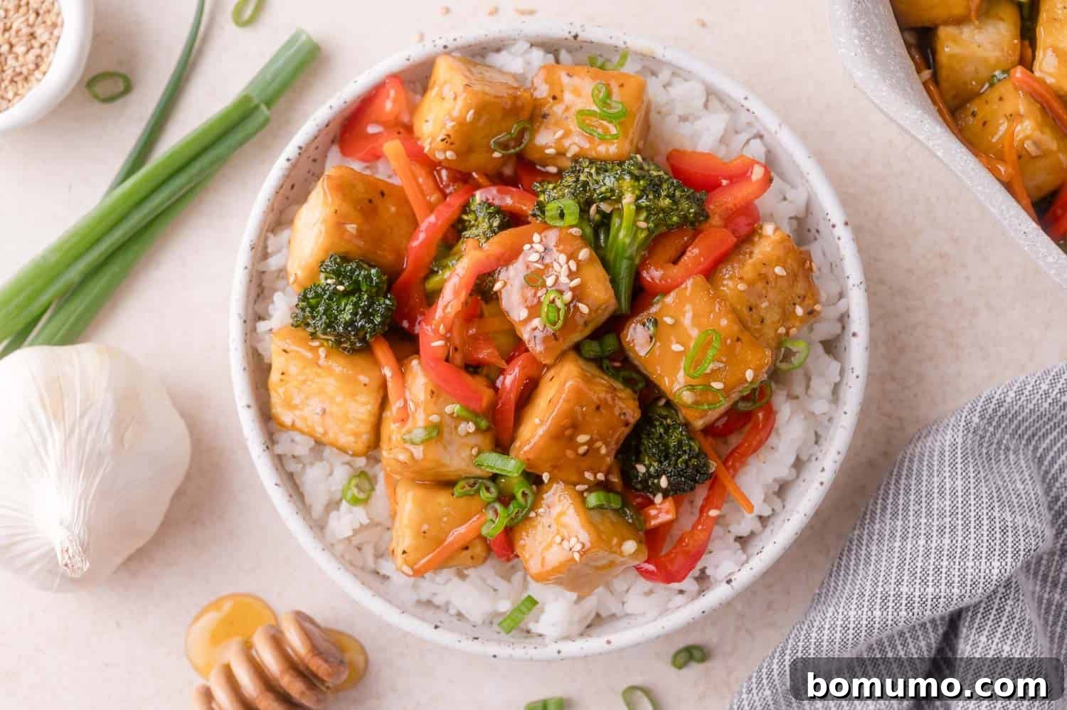 A bowl of rice topped with tofu cubes, red bell peppers, broccoli, sesame seeds, and chopped green onions, with a garlic bulb, scallions, and honey dipper nearby.