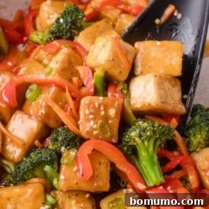 Stir-fried tofu cubes with broccoli and red bell pepper, garnished with sesame seeds and sliced green onions, served with a black spatula.