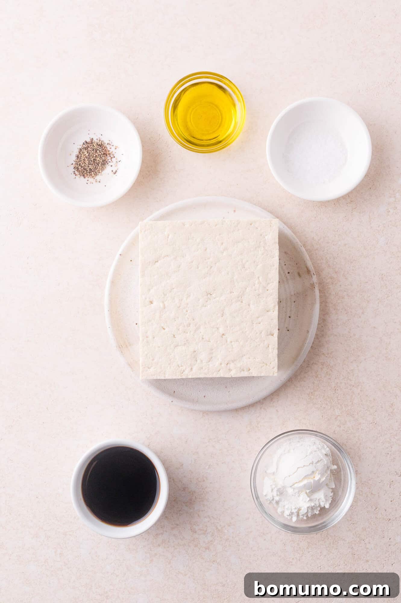 A block of tofu on a plate surrounded by small bowls containing black pepper, olive oil, salt, soy sauce, and cornstarch on a light surface.