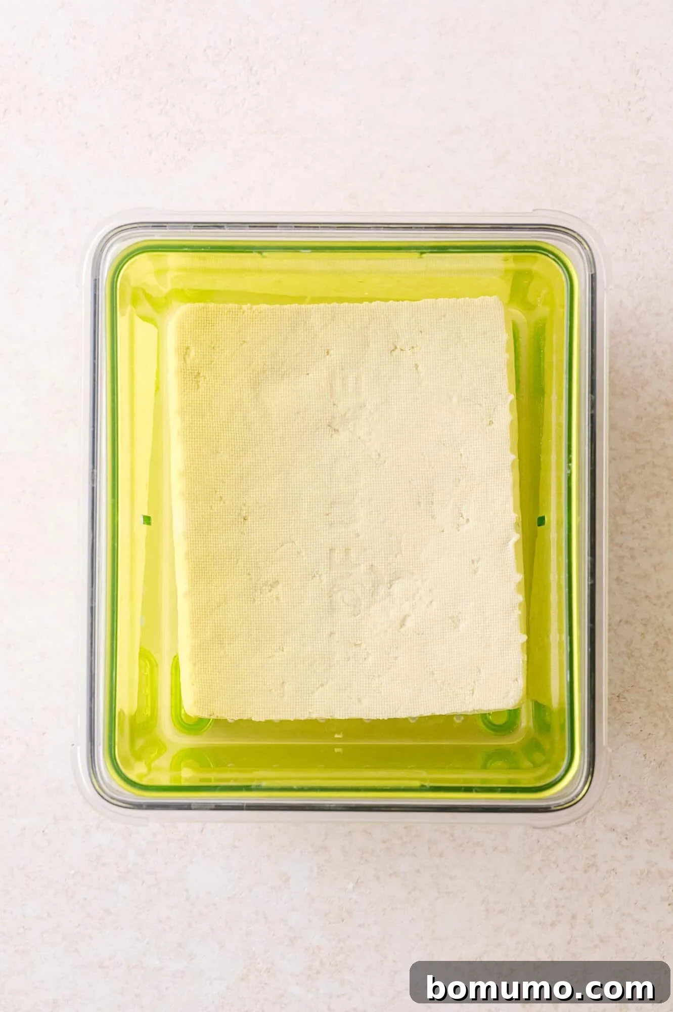 A block of tofu sits inside a green plastic container, viewed from above on a light-colored surface.