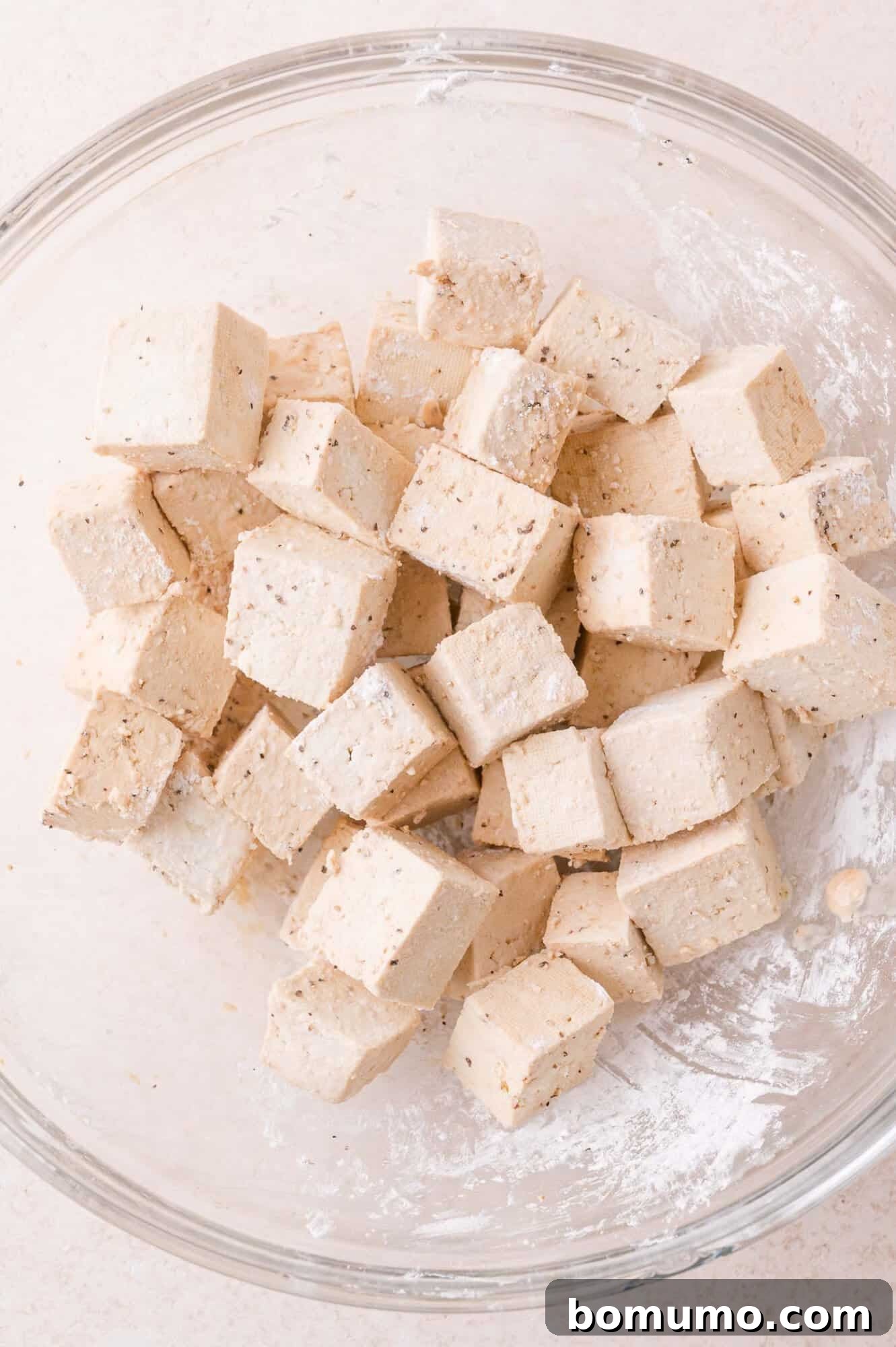 Cubed tofu coated with seasoning in a clear glass mixing bowl, set on a light-colored surface.