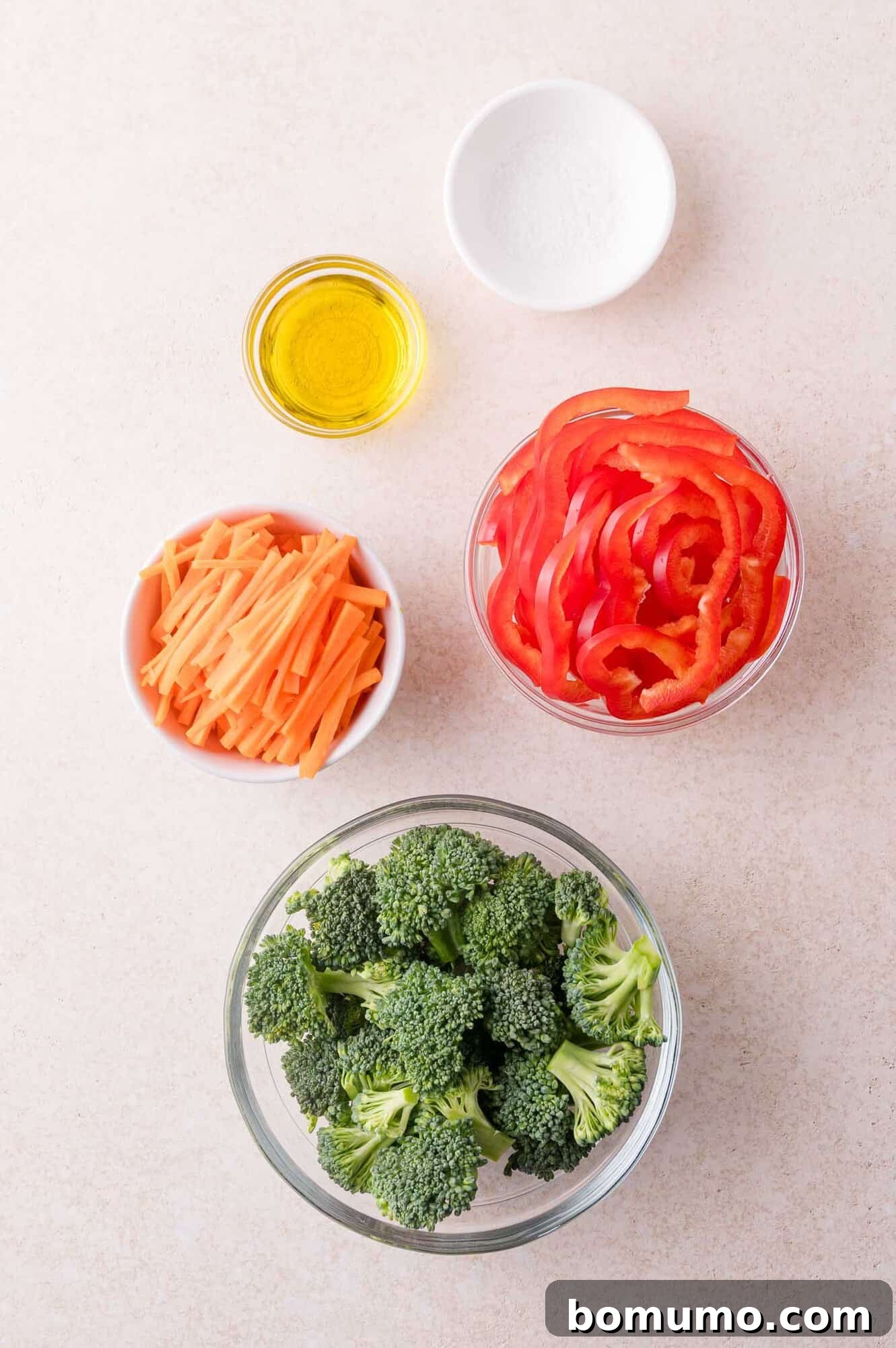 Glass bowls with broccoli florets, sliced red bell pepper, julienned carrots, a small bowl of olive oil, and a small bowl of salt on a light surface.