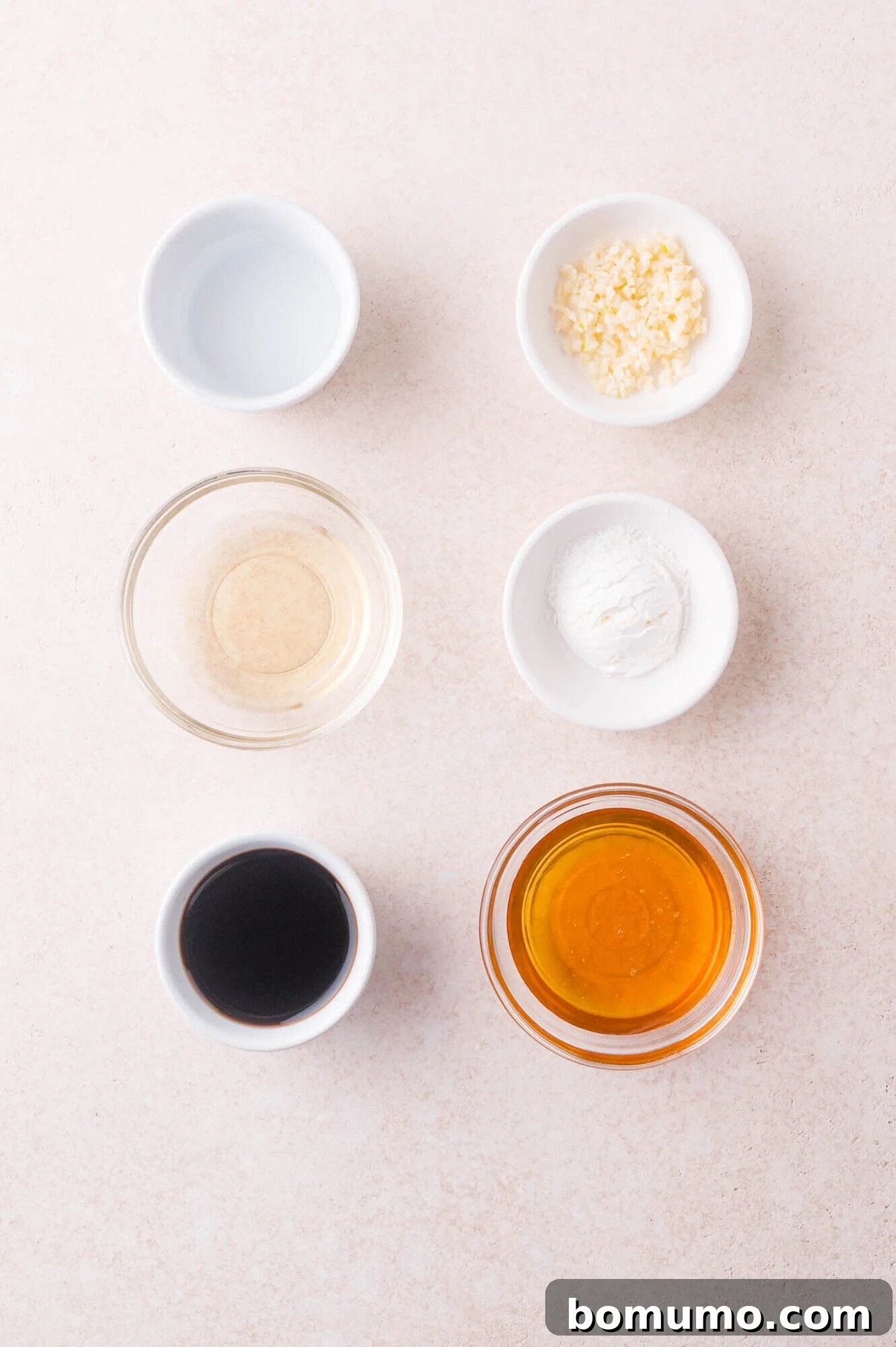 Six small bowls with various ingredients: chopped garlic, cornstarch, honey, soy sauce, rice vinegar, and water, arranged on a light-colored surface.