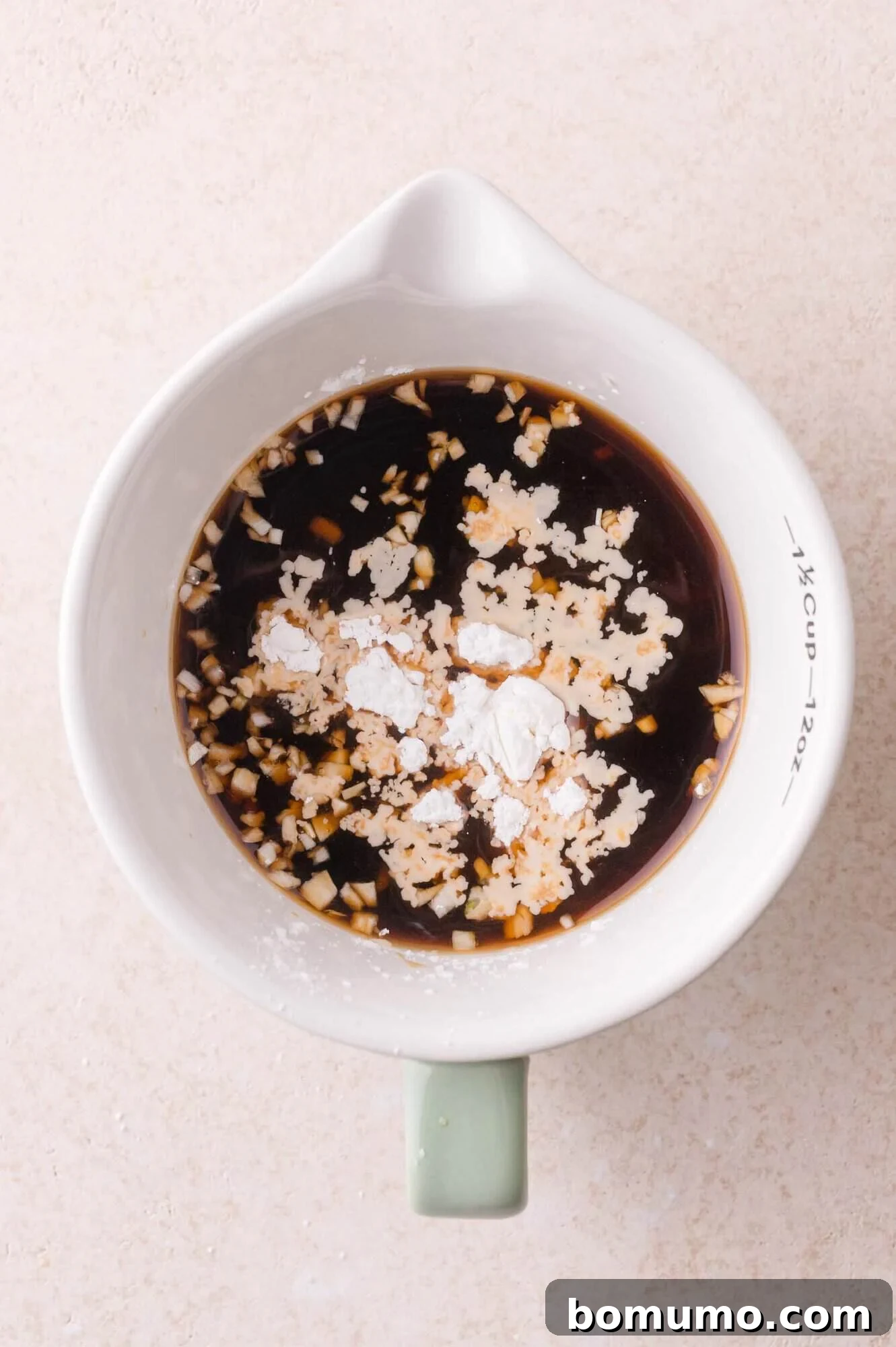 A white measuring cup with soy sauce, minced garlic, and cornstarch mixed together on a light countertop.
