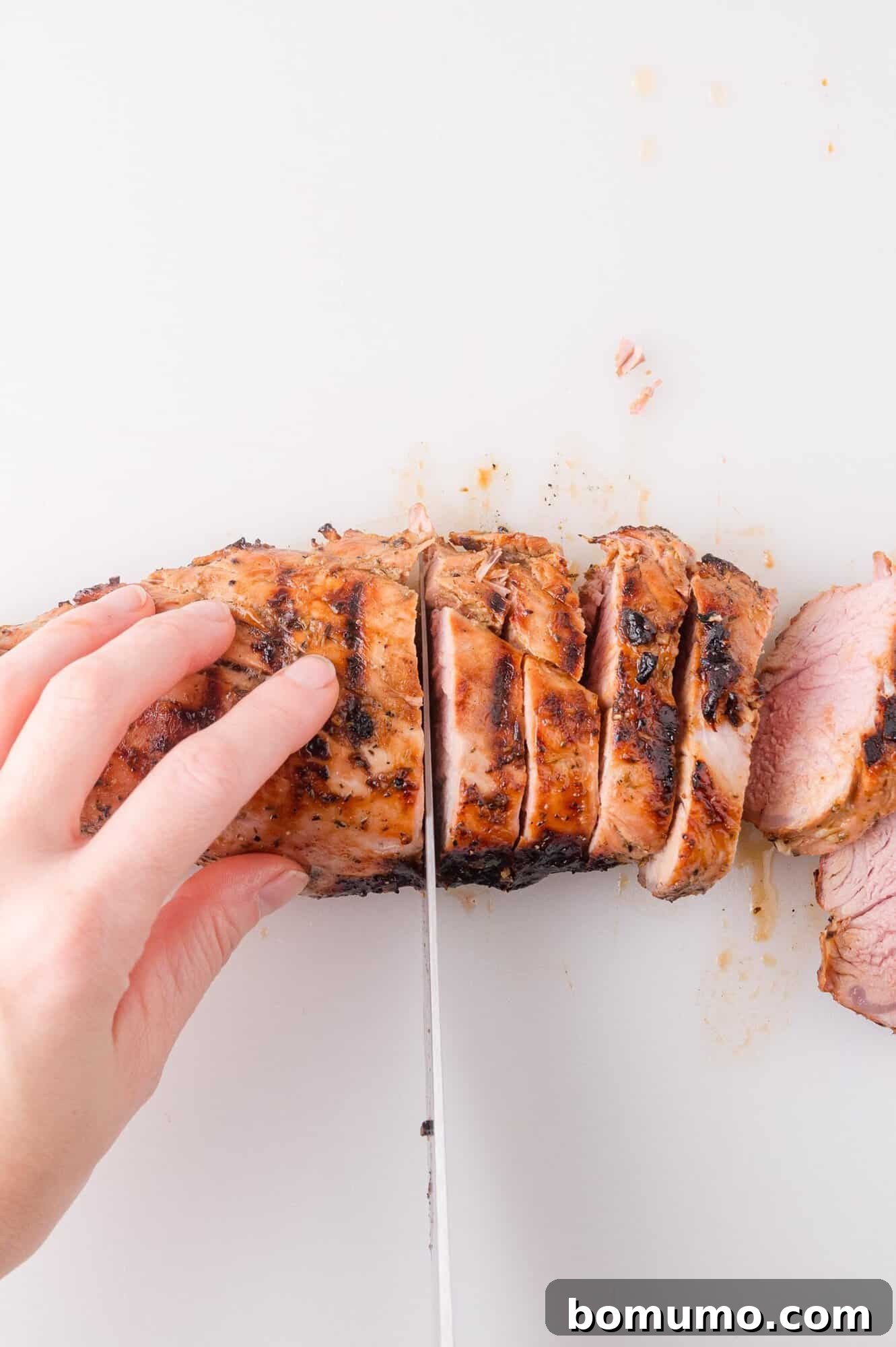 A hand slices cooked pork tenderloin into pieces on a white cutting board.