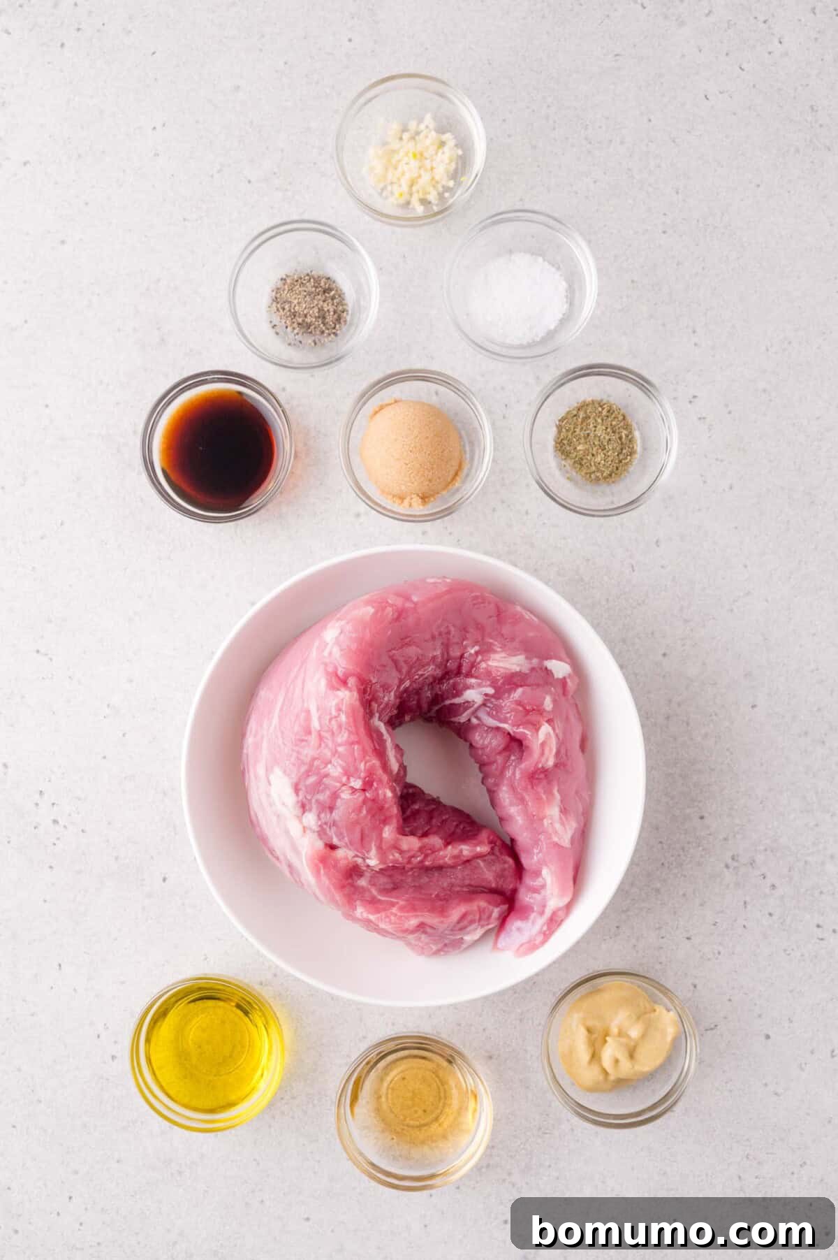 Raw pork tenderloin in a white bowl surrounded by small bowls of seasoning, oil, soy sauce, mustard, brown sugar, and minced garlic on a light surface.