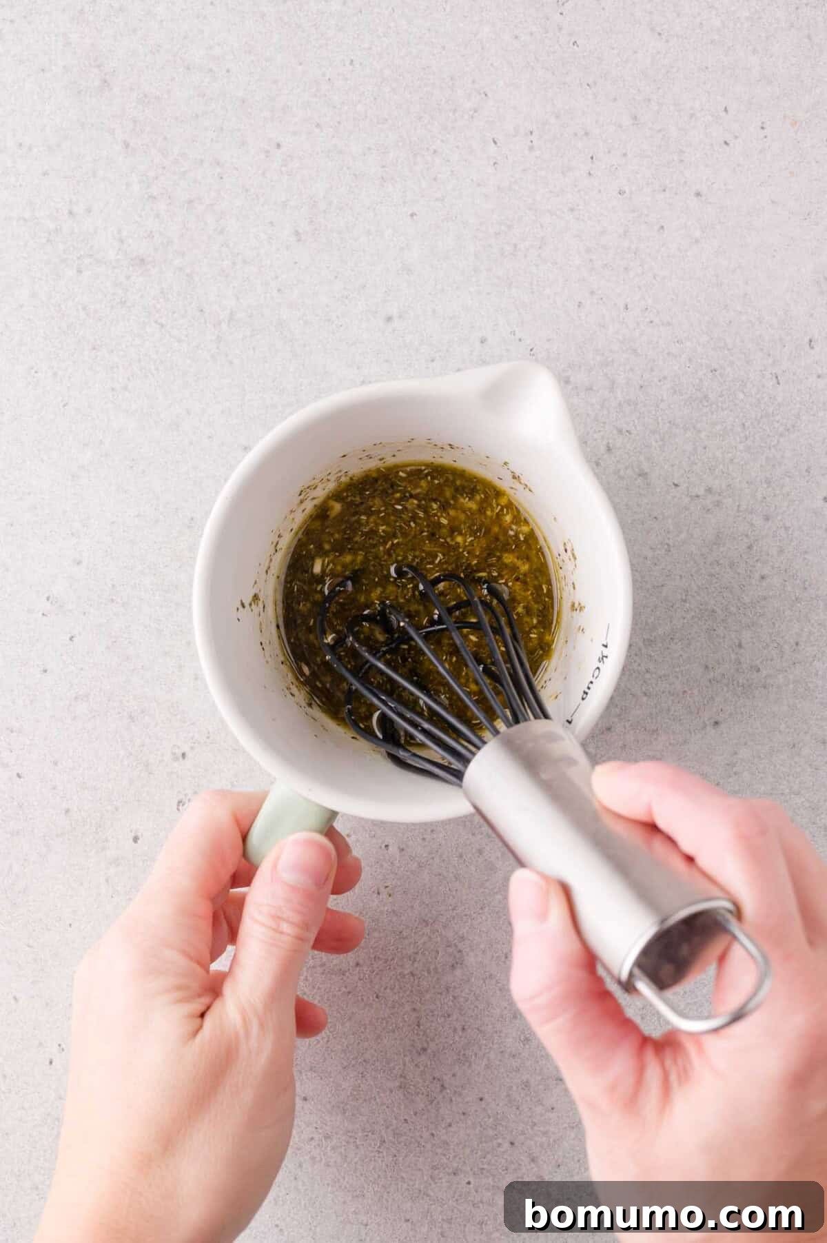 A person whisking marinade in a white ceramic measuring cup on a light gray surface.