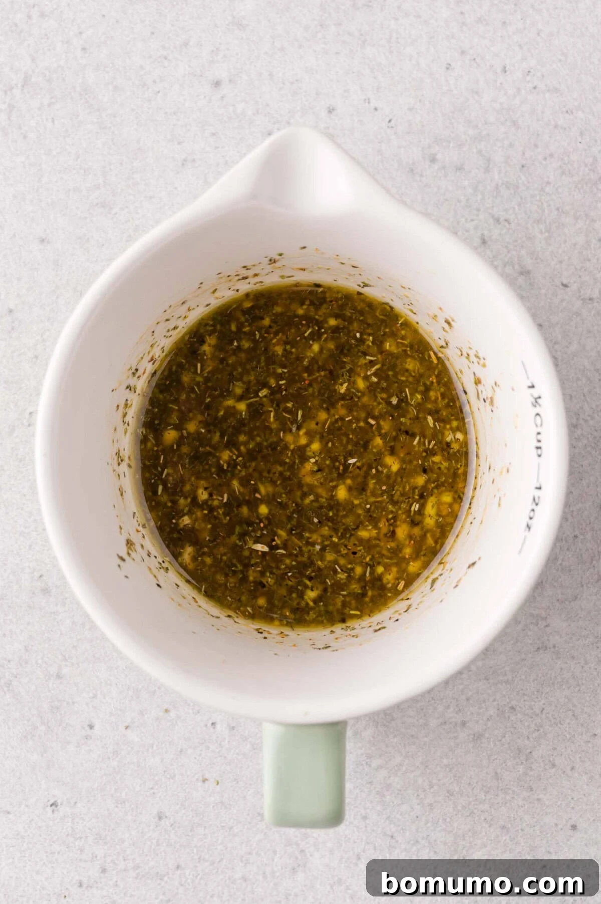 A white measuring cup filled with a mixed herb and oil marinade sits on a light gray surface.