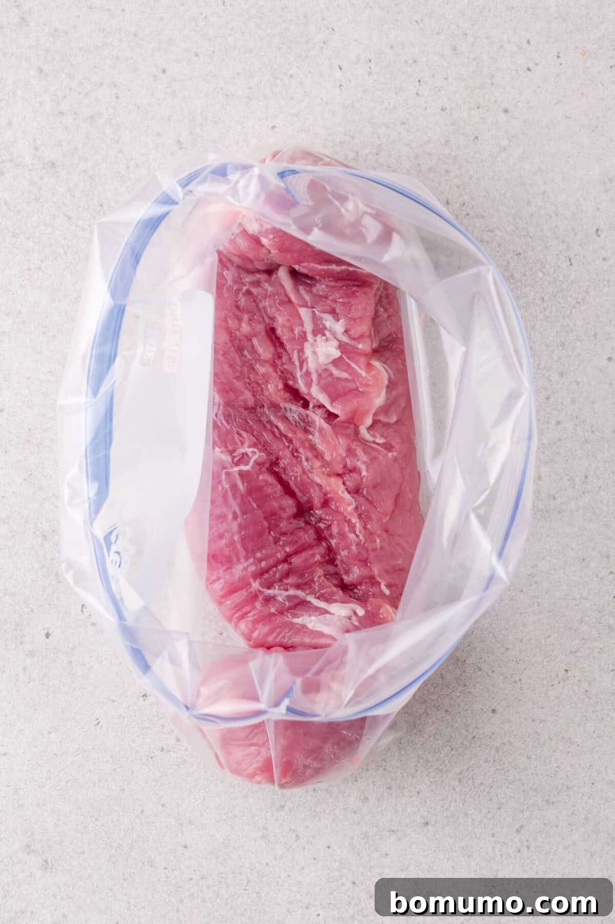 Raw pork tenderloin placed inside a clear resealable plastic bag, set on a light-colored surface.
